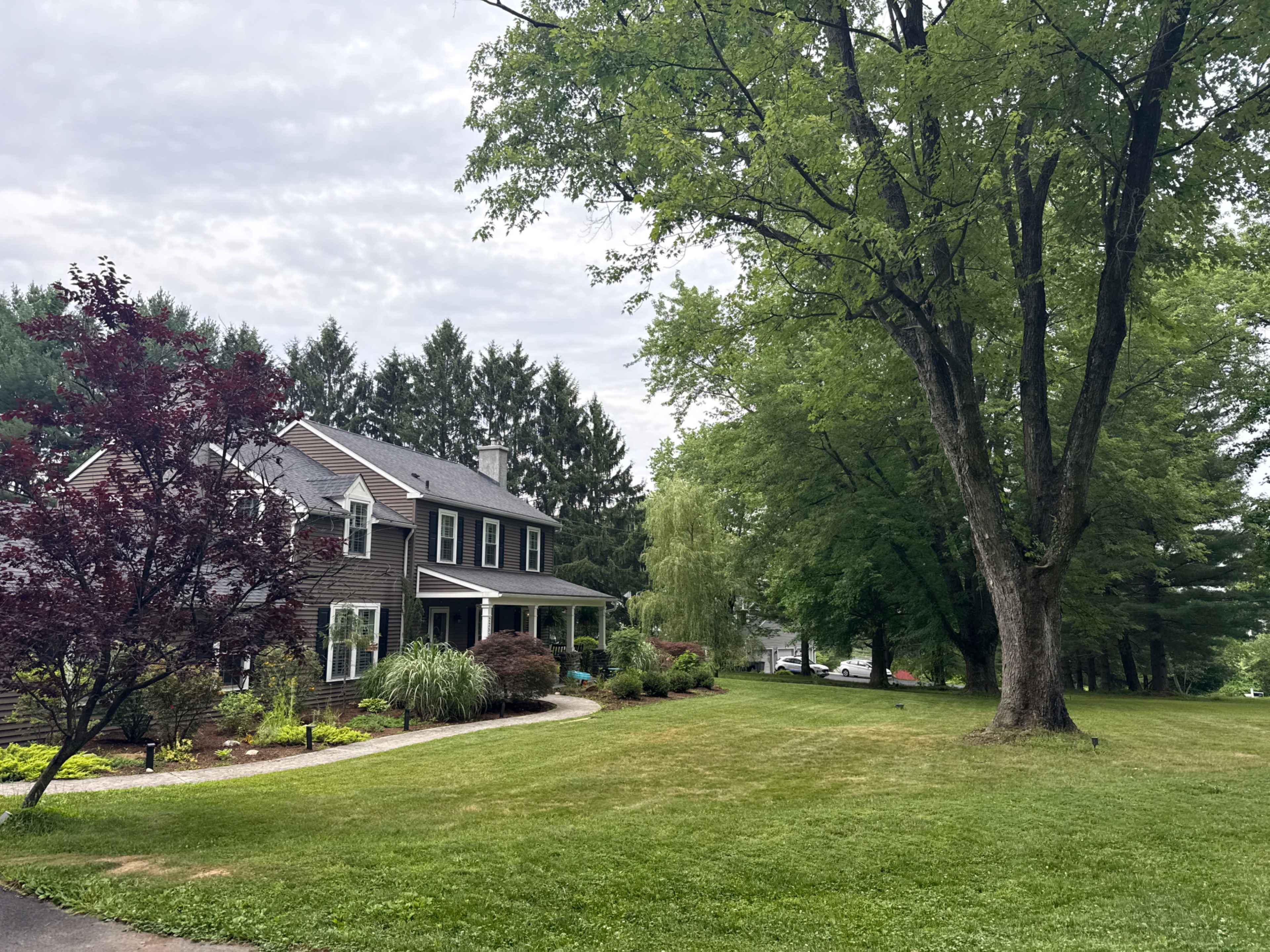 A two-story house with a dark exterior stands on a green lawn surrounded by trees and shrubs.