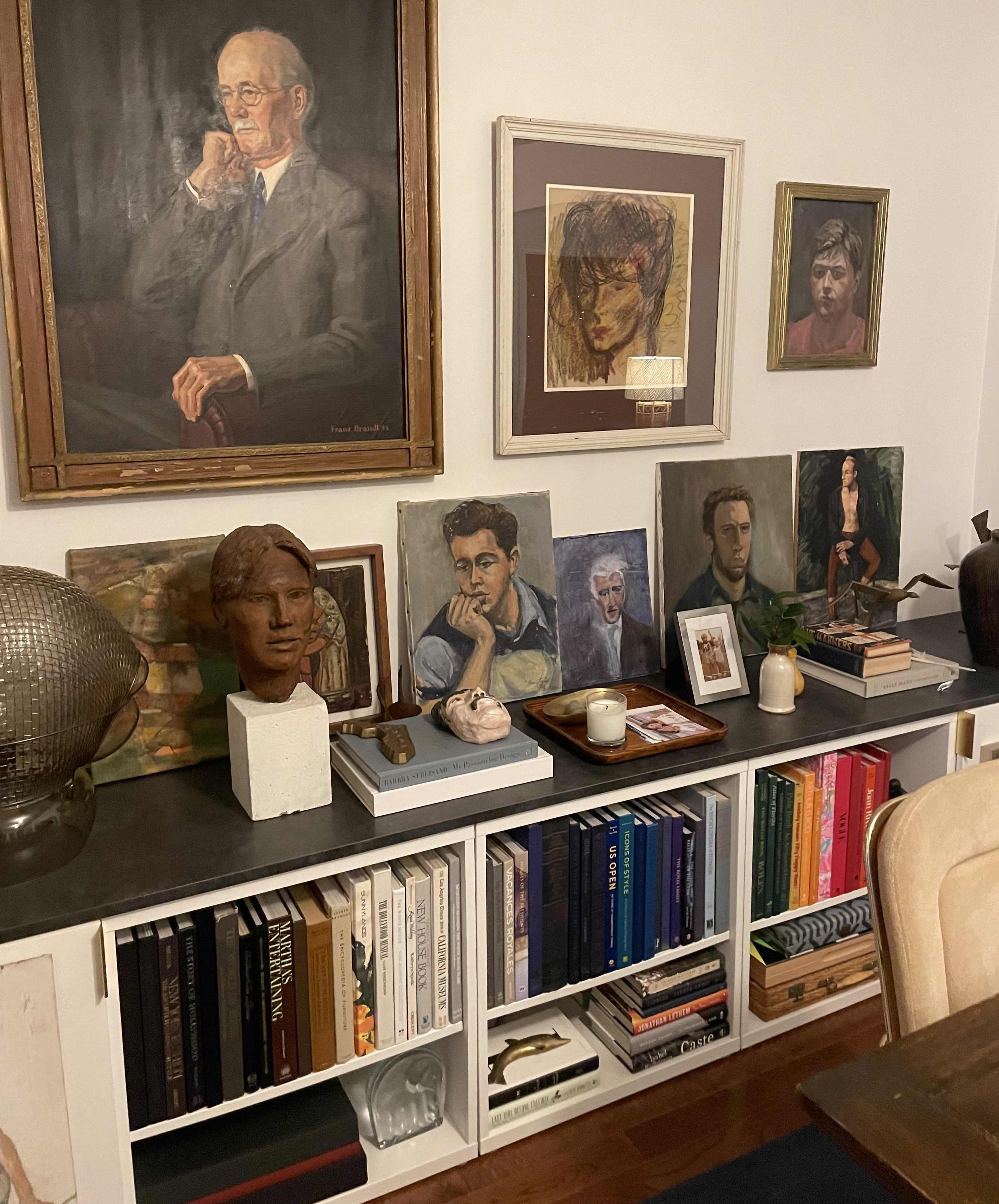 The image shows a bookshelf displaying various art pieces, sculptures, and a collection of books, with a large portrait hanging above the shelf.