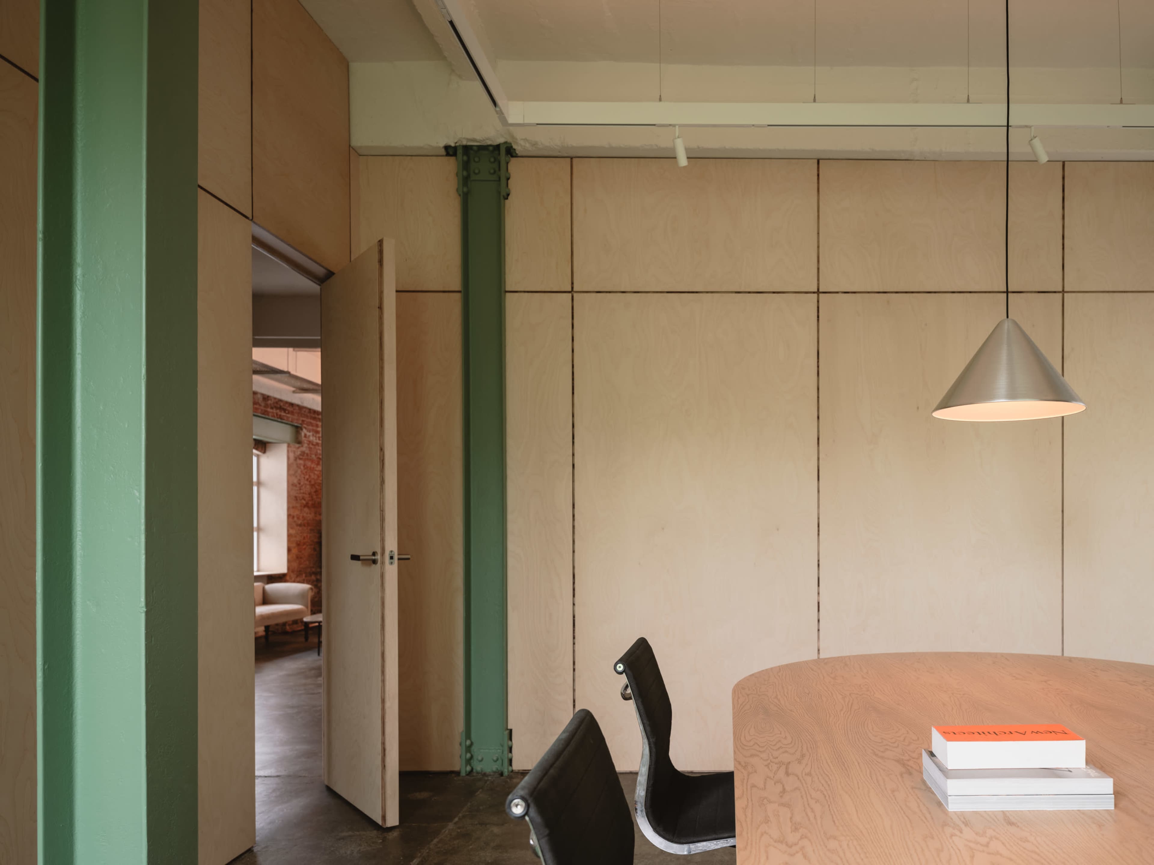 A modern meeting room features a round wooden table, sleek chairs, and a pendant light, with green beams and wood-paneled walls.