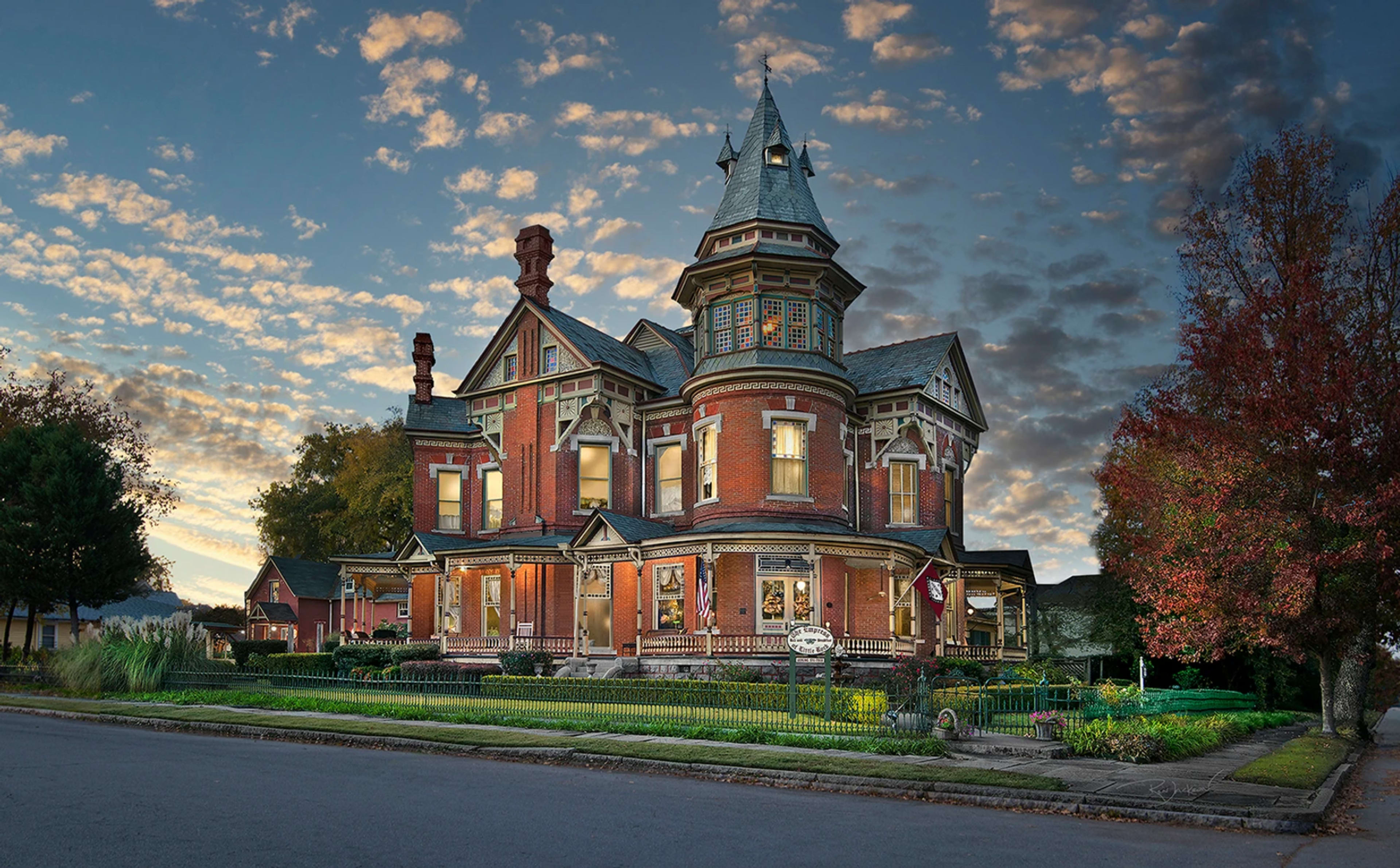 The image depicts a large, ornate Victorian mansion with a tall turret, surrounded by a well-kept garden and a fence, set against a backdrop of a colorful sky at dusk.