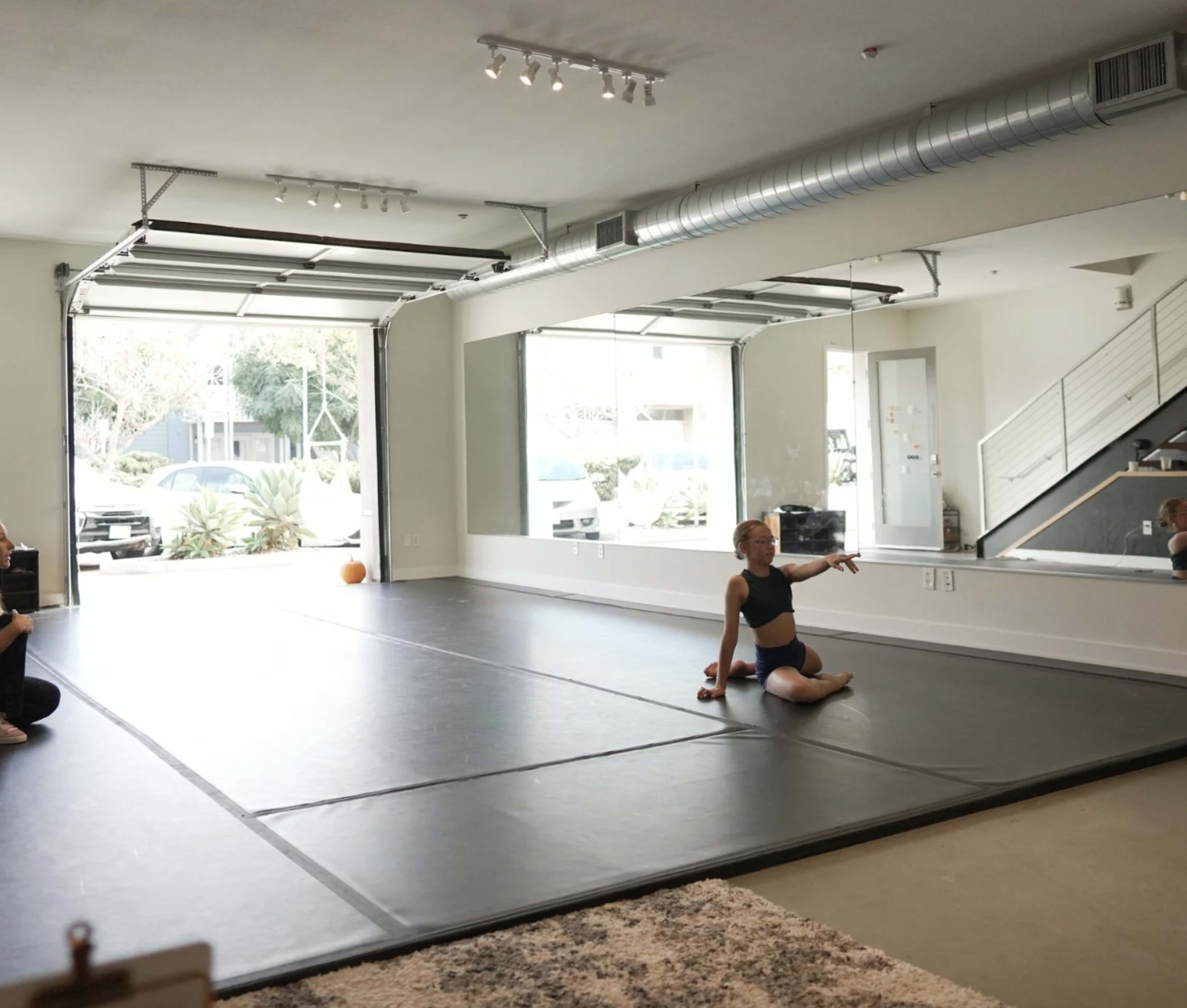 A dancer performs on a black mat in a spacious studio with large mirrors and an open garage door.
