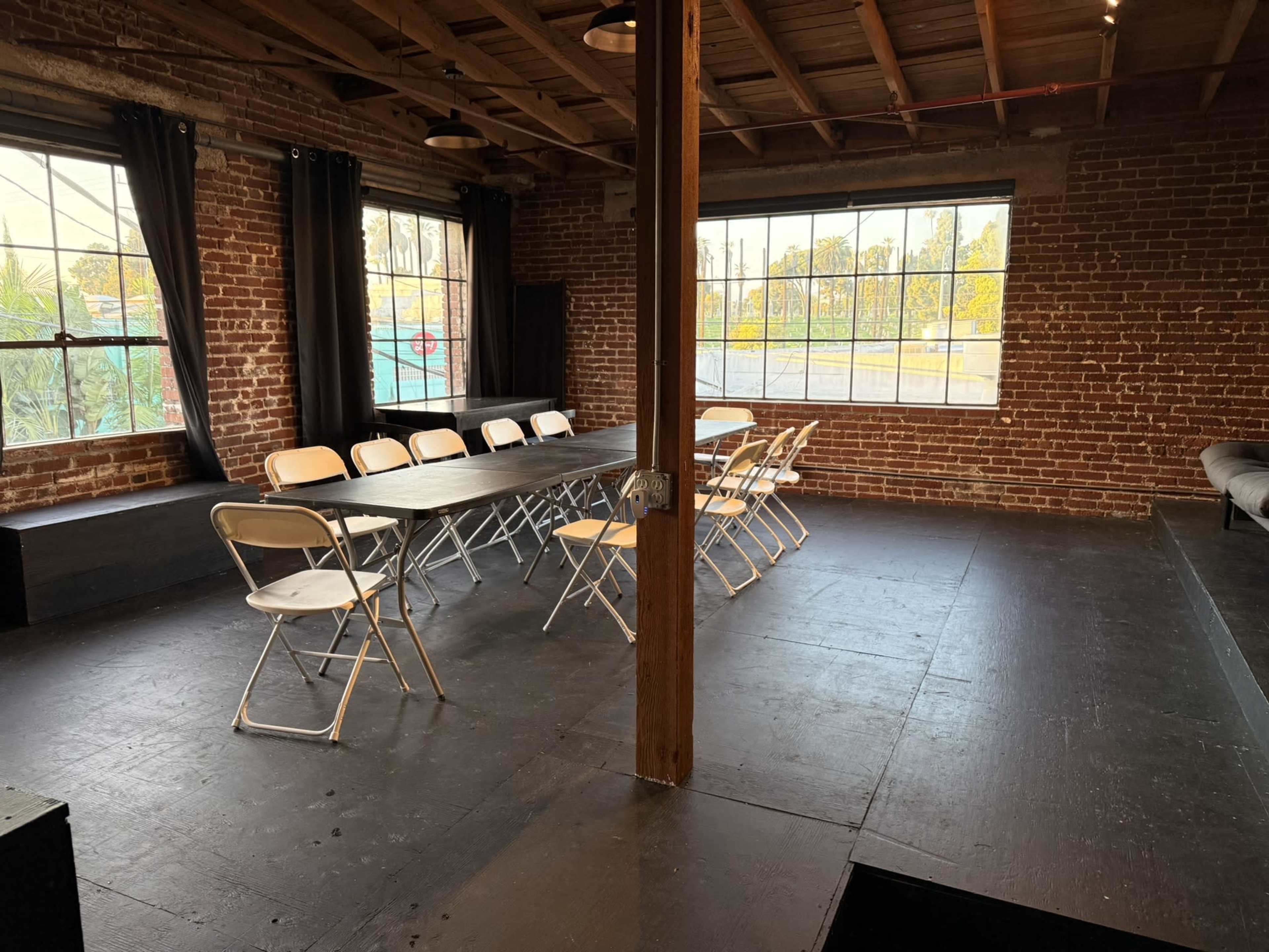 The image shows a spacious room with exposed brick walls, a large table surrounded by folding chairs, and large windows allowing natural light to enter.