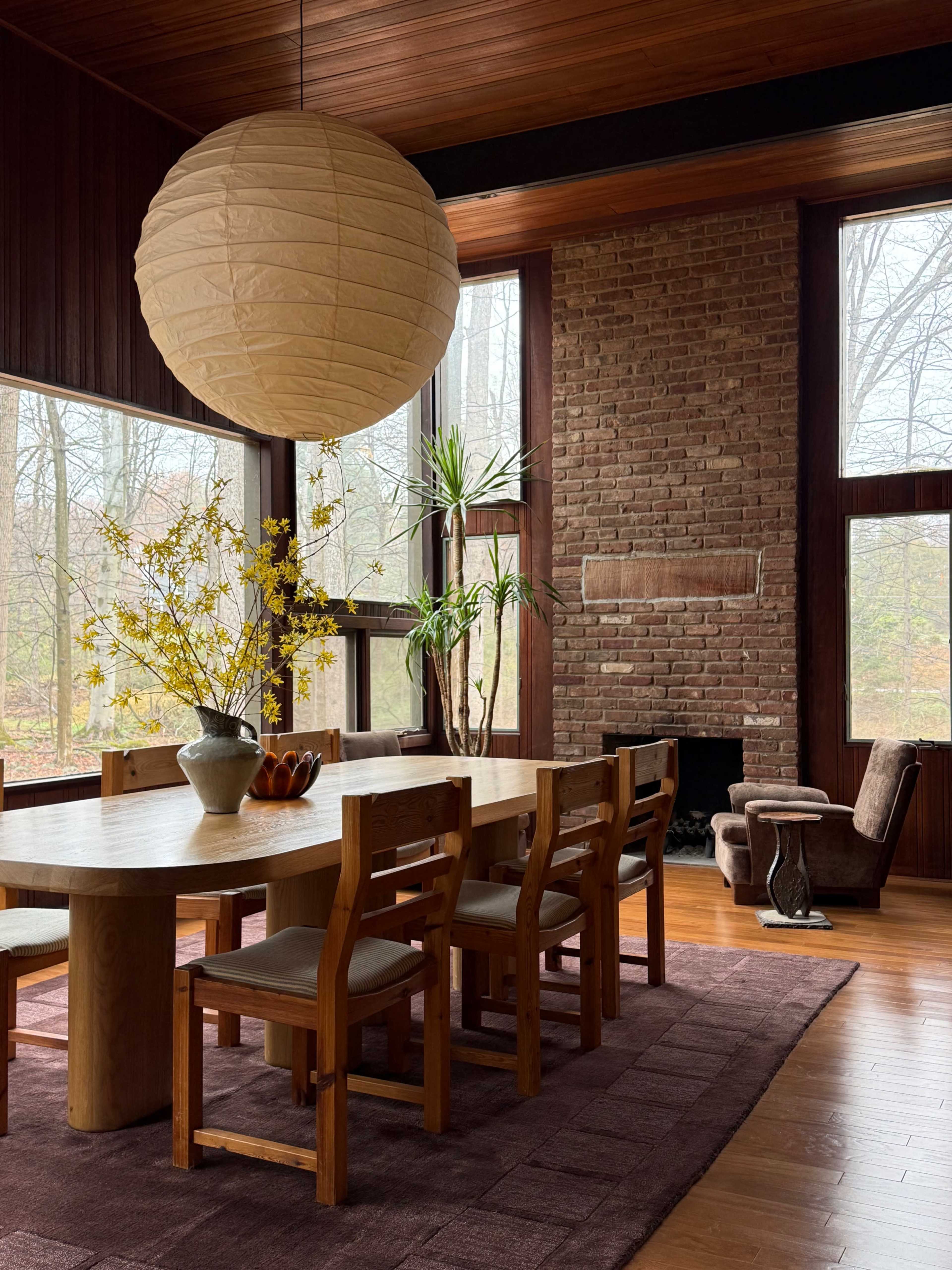 A dining area features a large wooden table surrounded by chairs, a vase with yellow flowers, and large windows that overlook a wooded area, with a brick fireplace in the background.