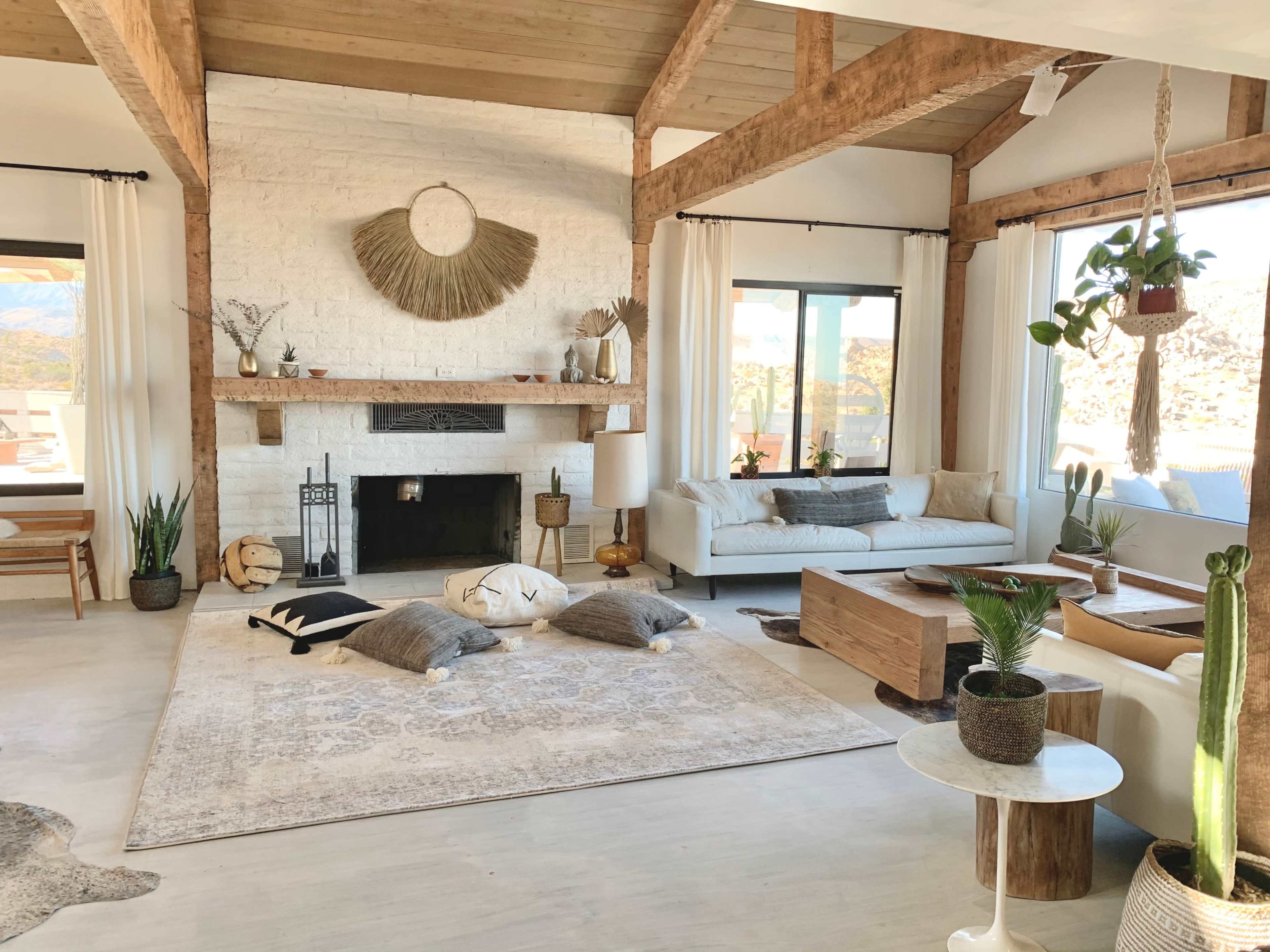 An airy living room with a stone fireplace, wooden beams, large windows, a plush area rug, and various plants scattered throughout the space.