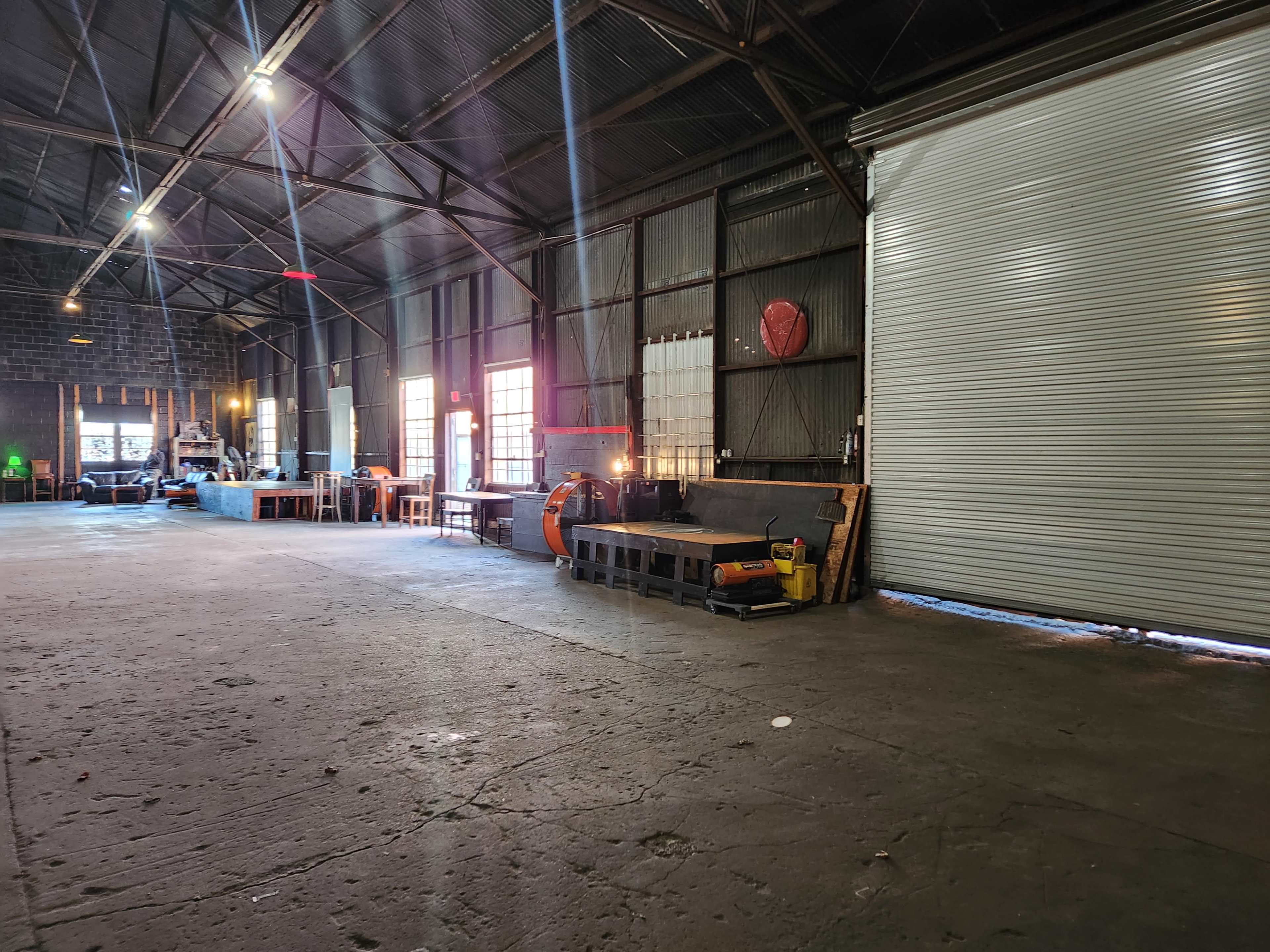 A spacious industrial warehouse with benches, tools, and a large rolled-up door on one side.