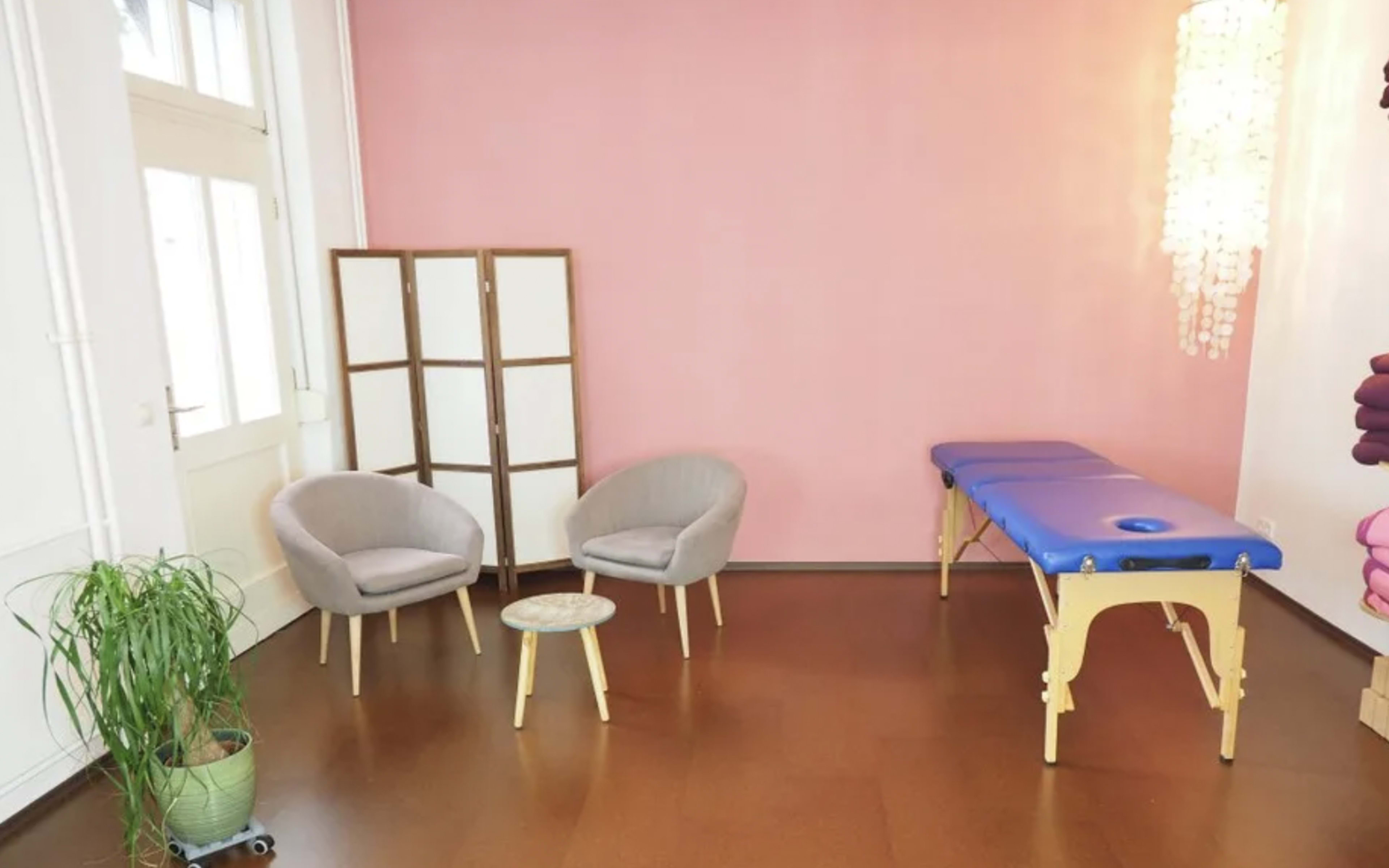A spacious room with a pink wall, featuring two gray chairs, a small table, and a massage table.