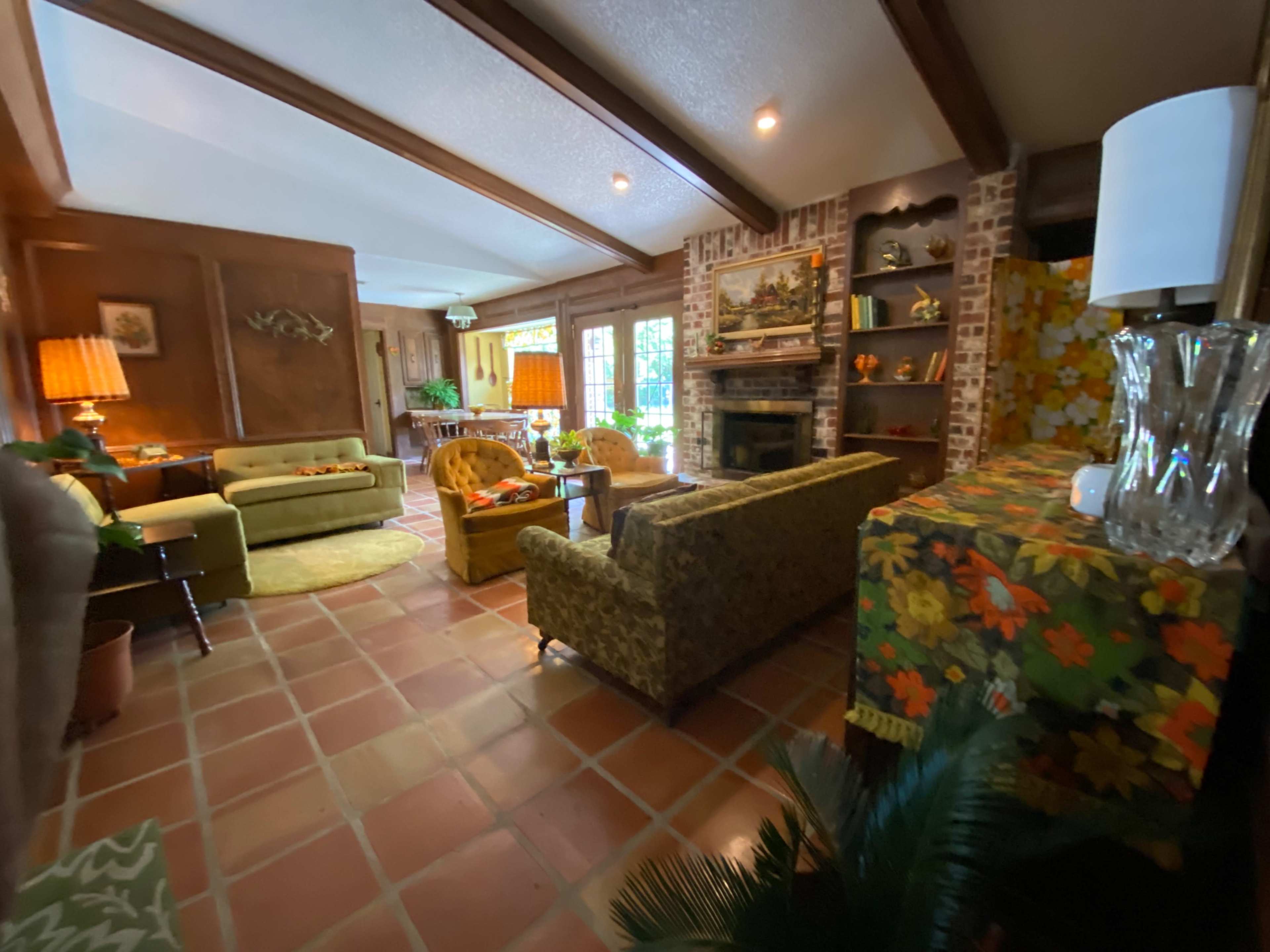 A cozy living room with a brick fireplace, patterned furniture, and large windows that allow natural light to enter.