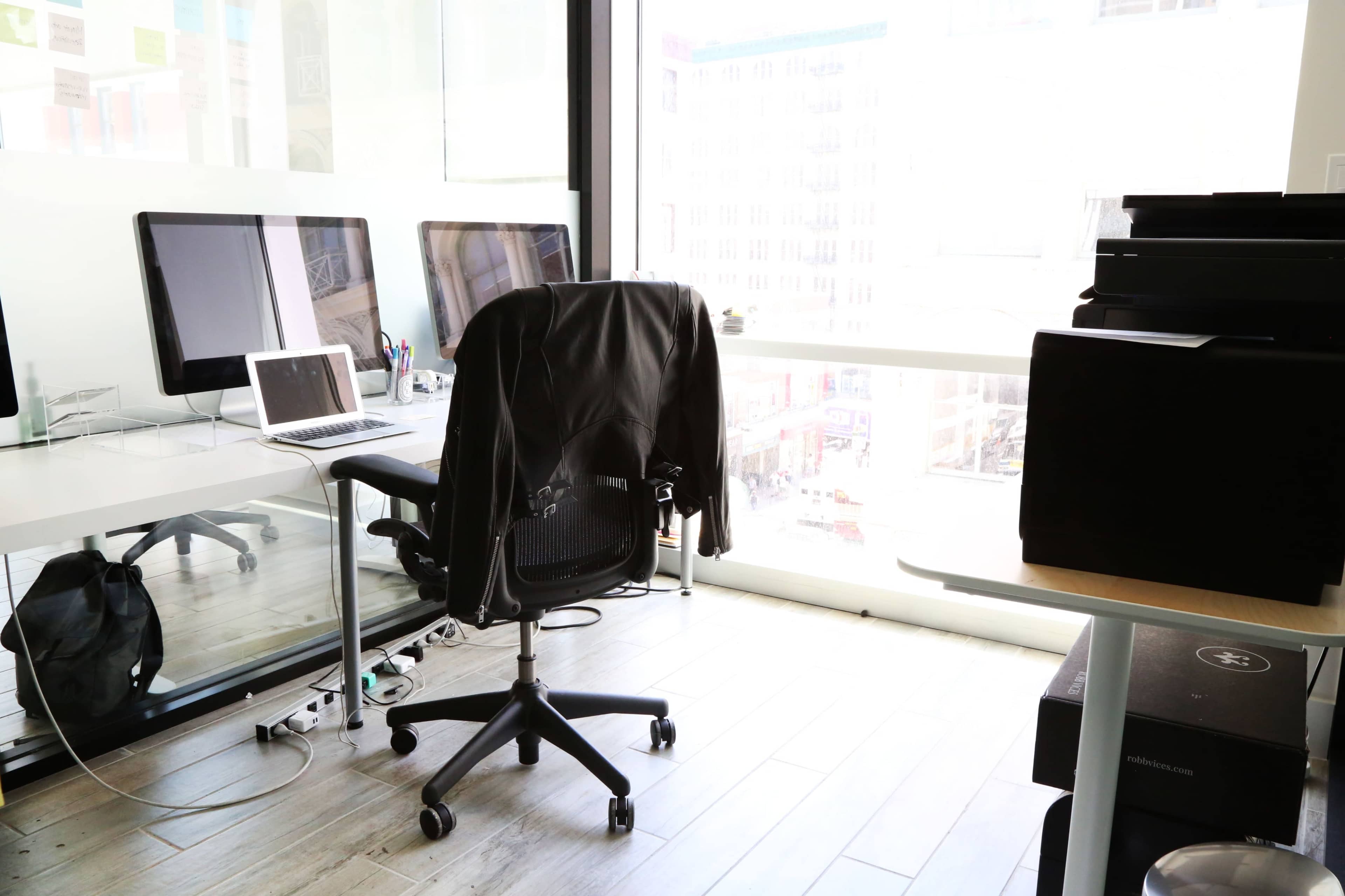 A modern office space with a desk, a swivel chair, two computer monitors, and a view of the city through large windows.