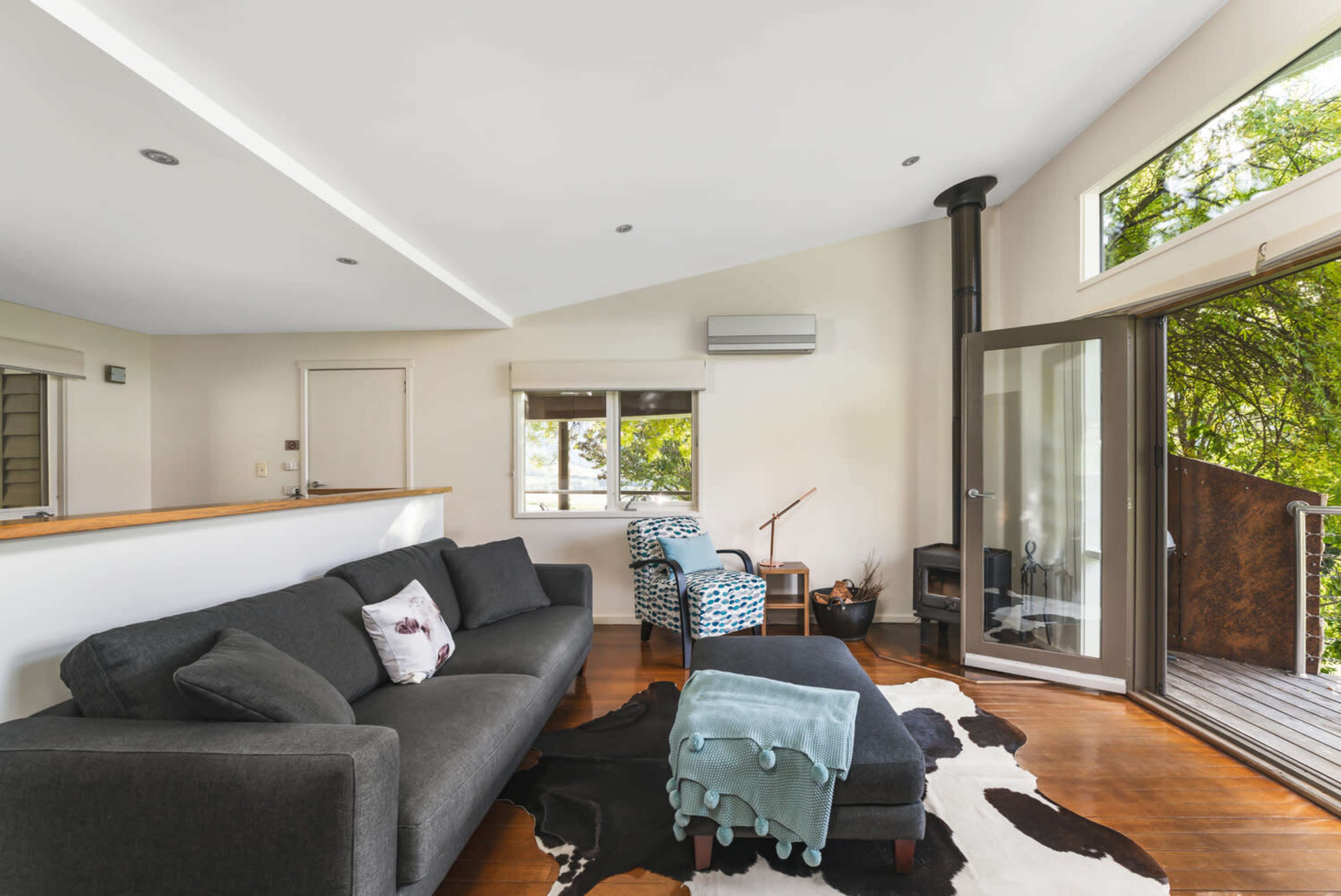 A cozy living room features a dark sofa, a patterned chair, a small table, and large windows opening to a view of greenery.