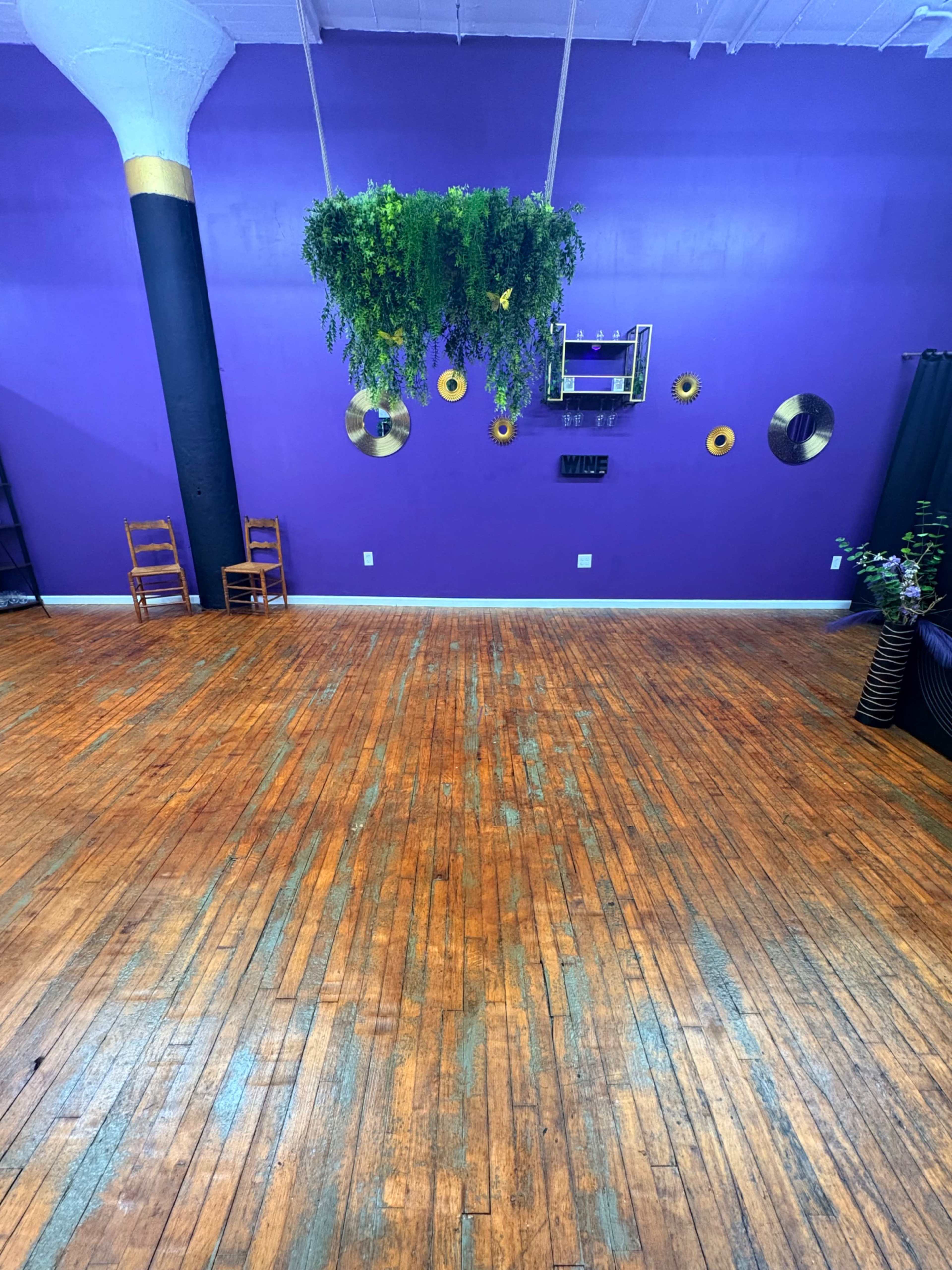The image shows a brightly colored purple studio space with wooden flooring, a hanging plant installation, and decorative items on the wall.