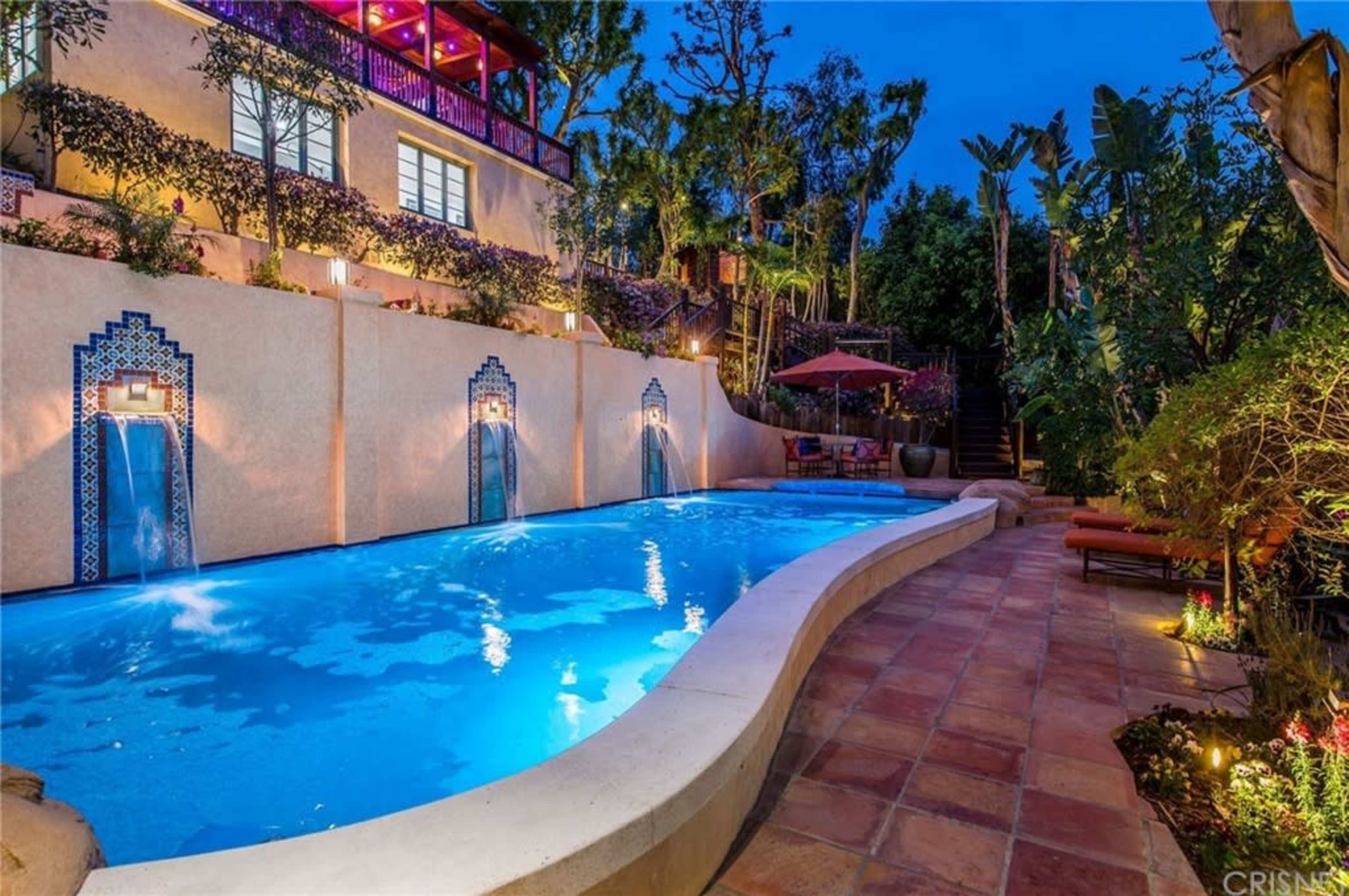 A curved swimming pool with three wall fountains is surrounded by lush greenery and outdoor seating, illuminated by soft lights at dusk.