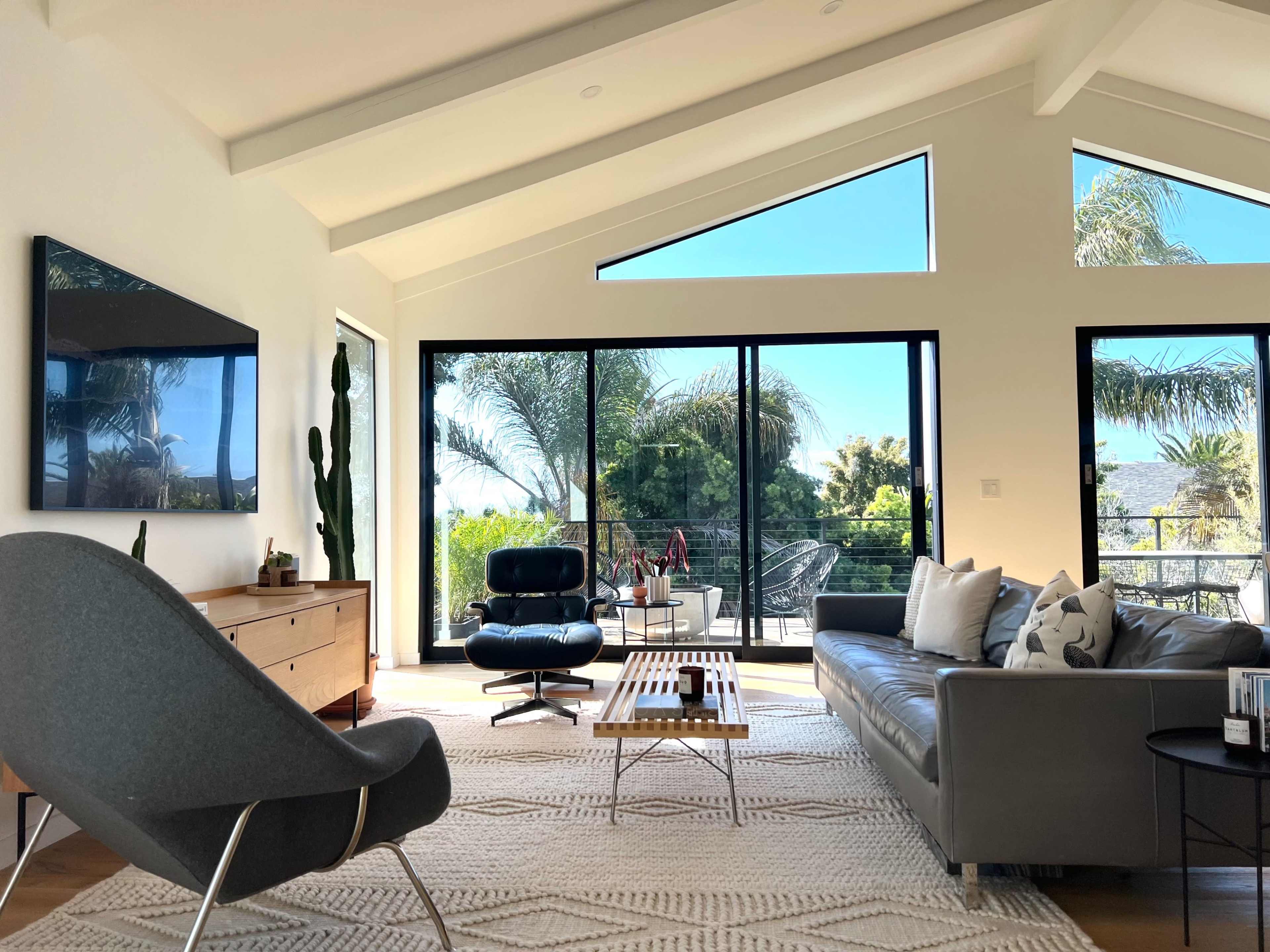 A modern living room with large windows overlooking a palm tree landscape, featuring a sectional sofa, an accent chair, a wooden coffee table, and a sleek television mounted on the wall.