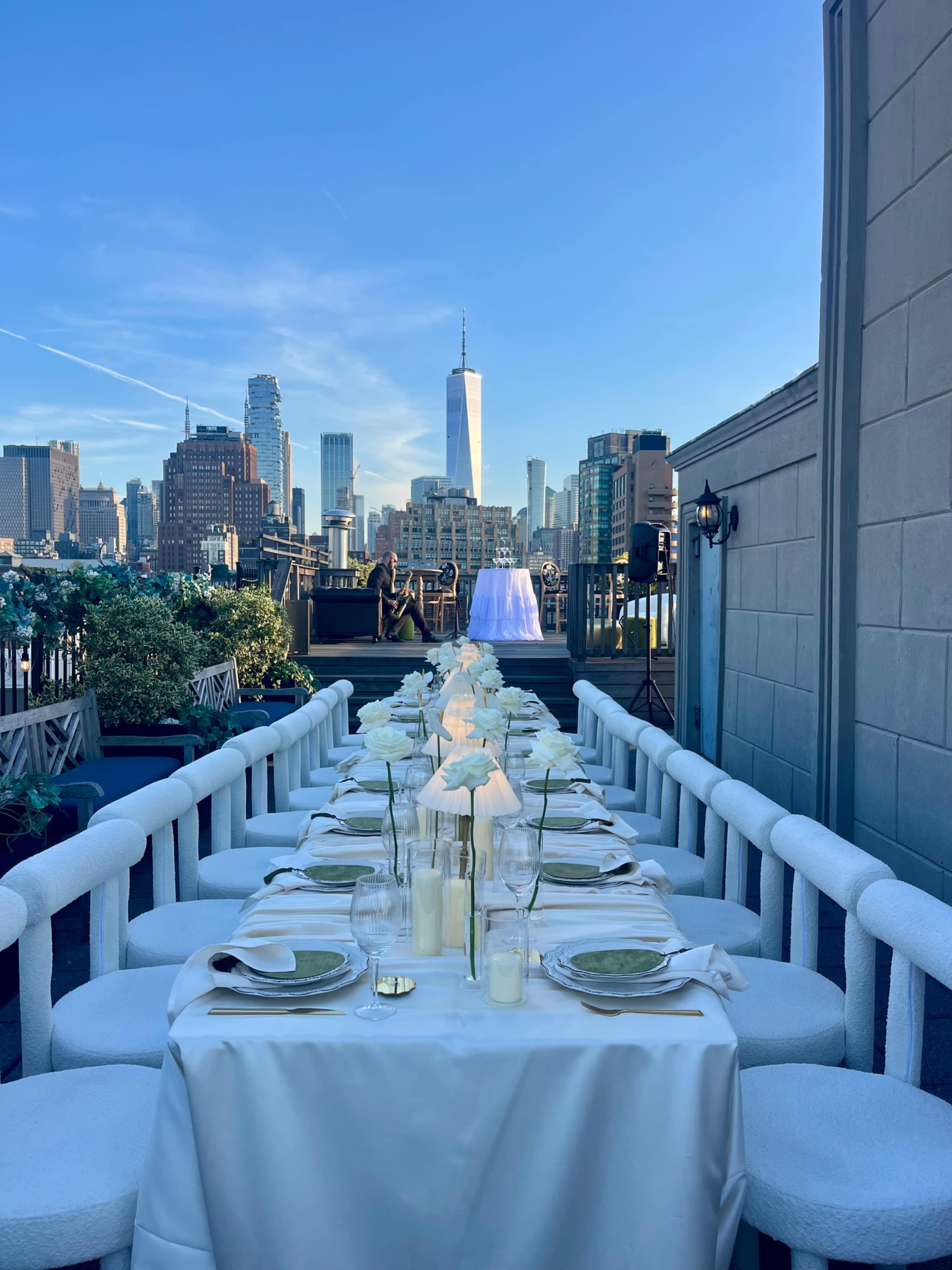 A long dining table set for a formal event overlooks a city skyline featuring One World Trade Center under a clear blue sky.