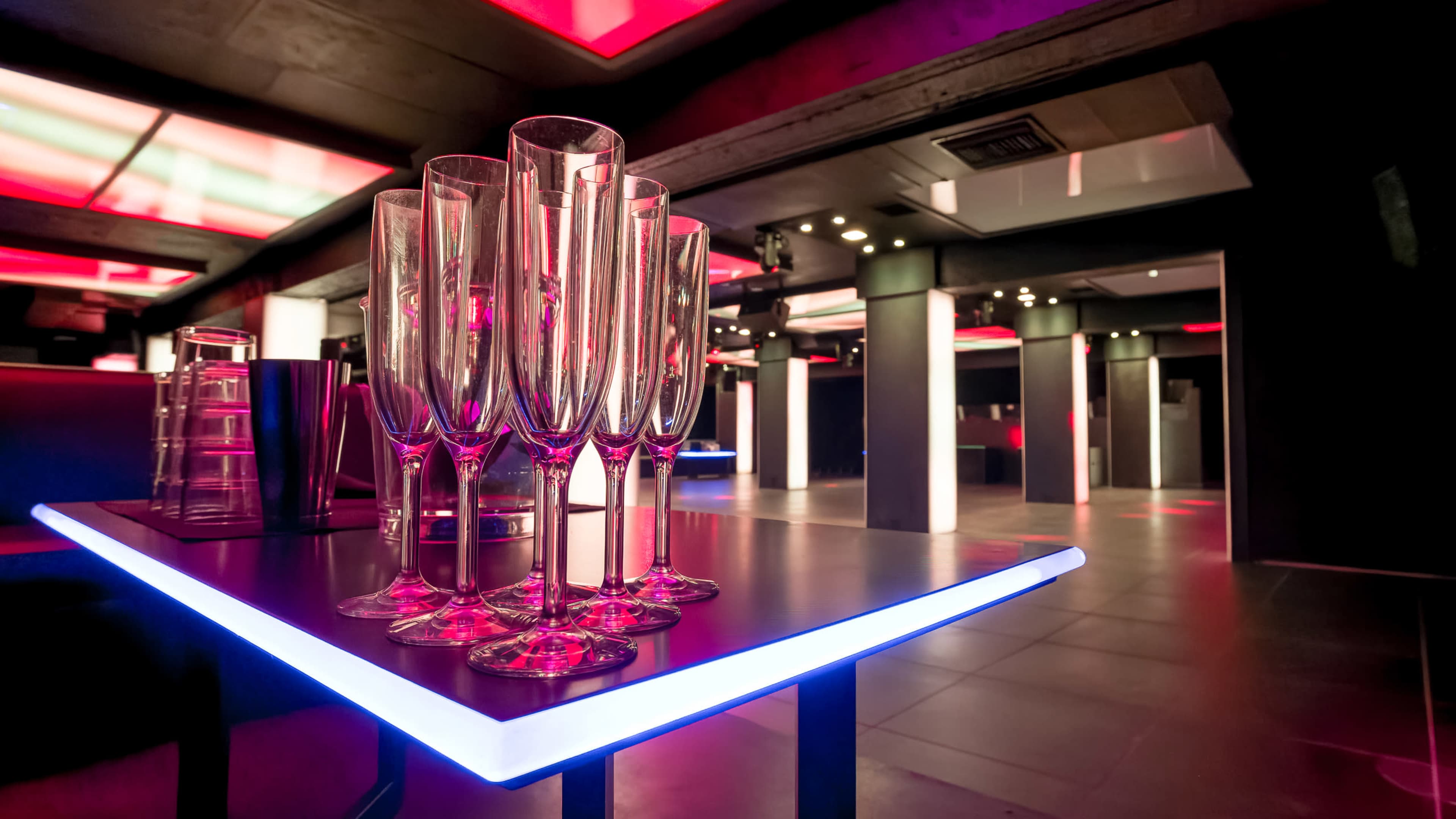 A set of champagne flutes is arranged on a illuminated table in a modern, dimly lit lounge featuring red and purple lighting.