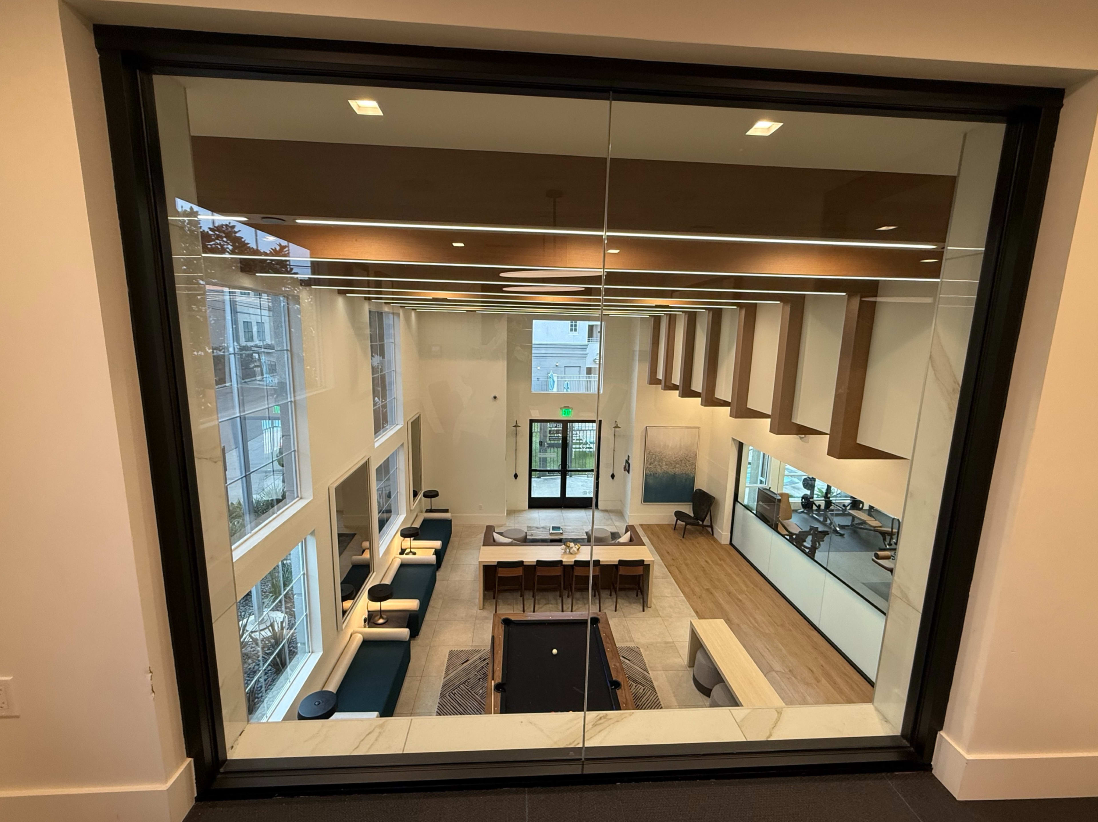 The image shows a spacious interior of a modern lounge or community room, featuring large windows, a dining area, and a pool table.