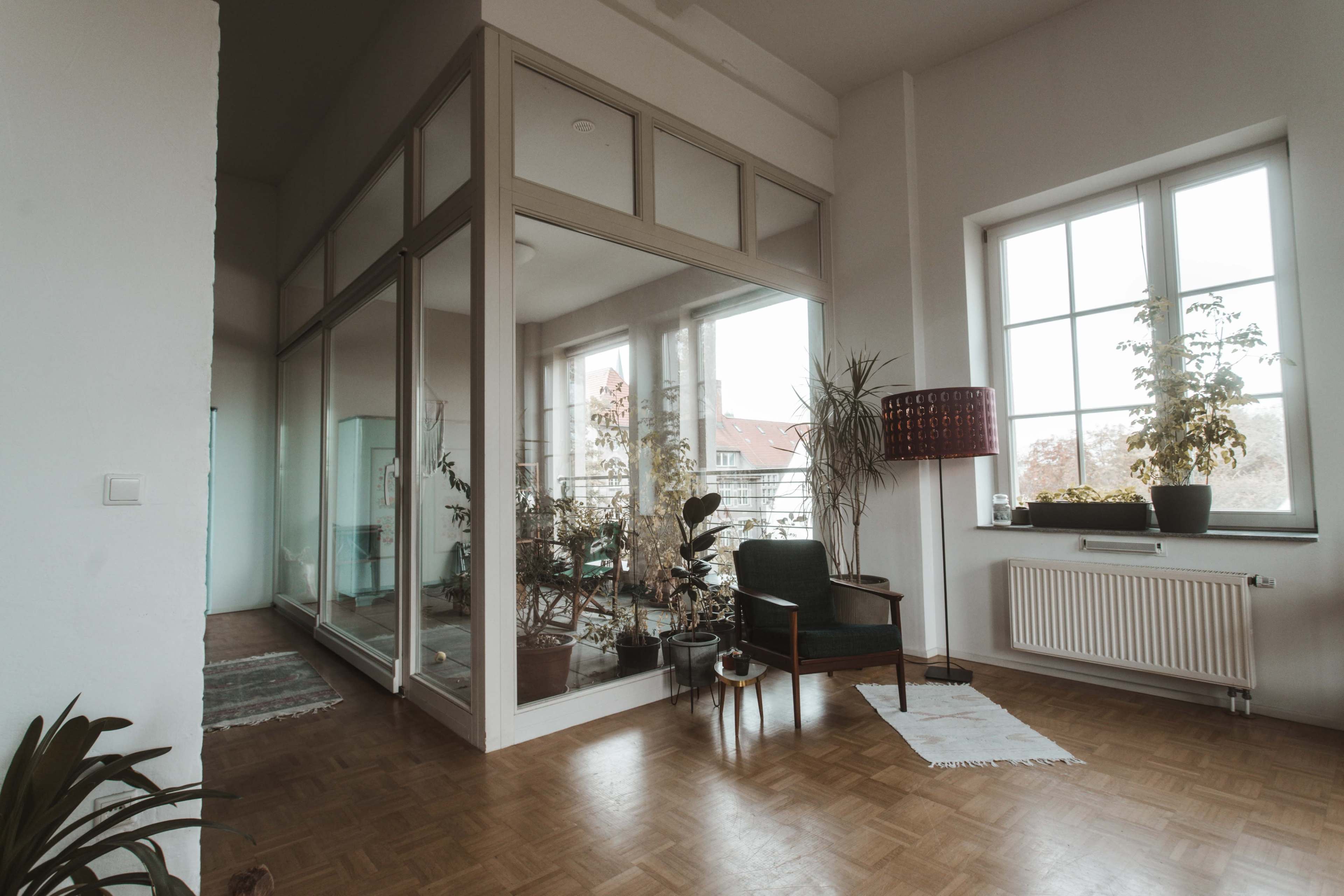 The image shows a bright room featuring a large glass-walled area with plants, a chair, and a radiator beneath a window.