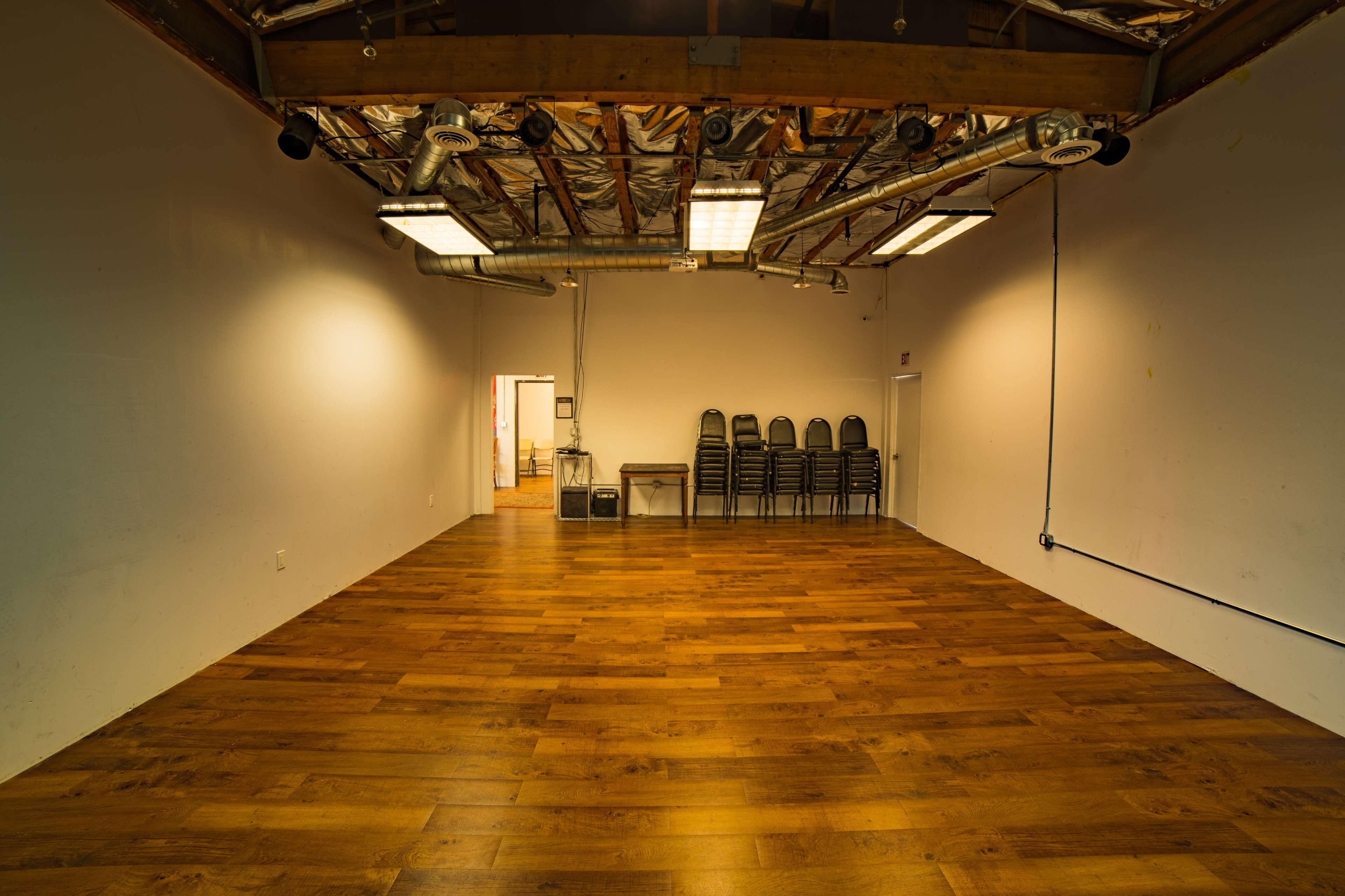 An empty room with wooden flooring, a stack of chairs against one wall, and exposed ceiling beams and fixtures.