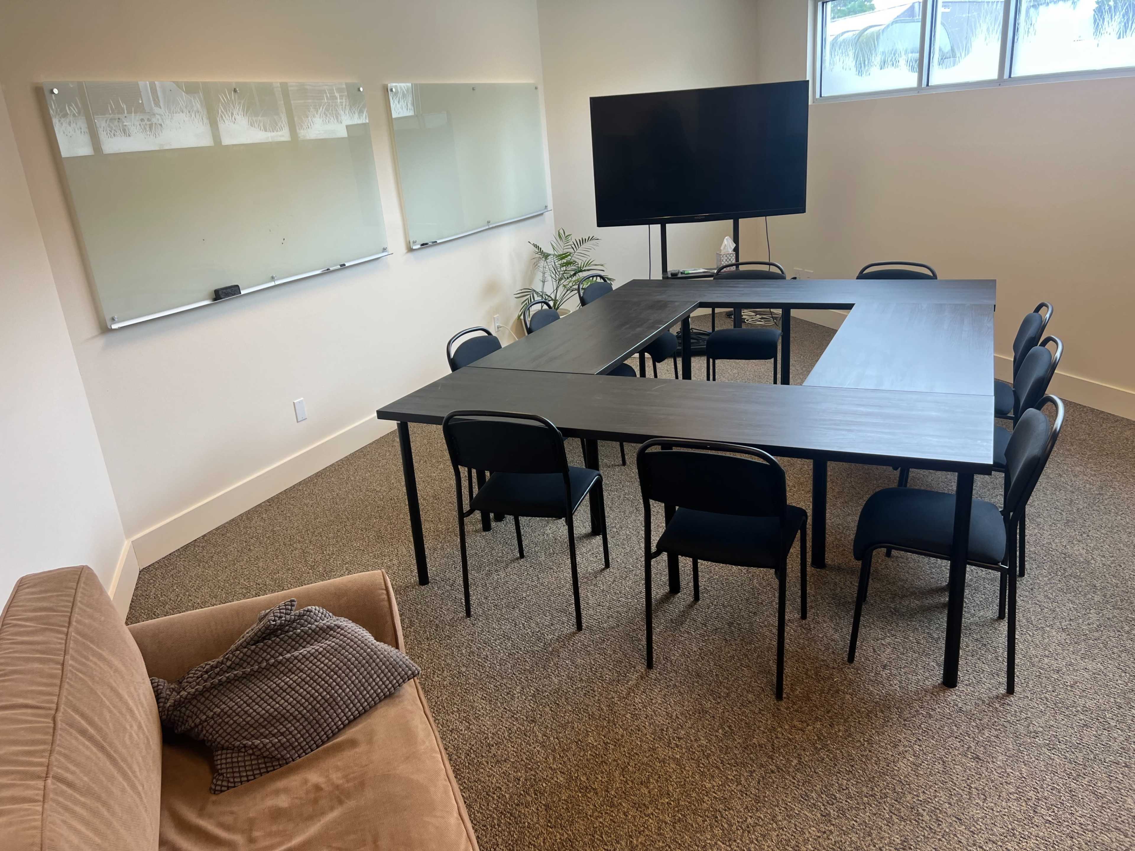 A meeting room features a large rectangular table surrounded by ten black chairs, with a couch in the corner and a television mounted on the wall.