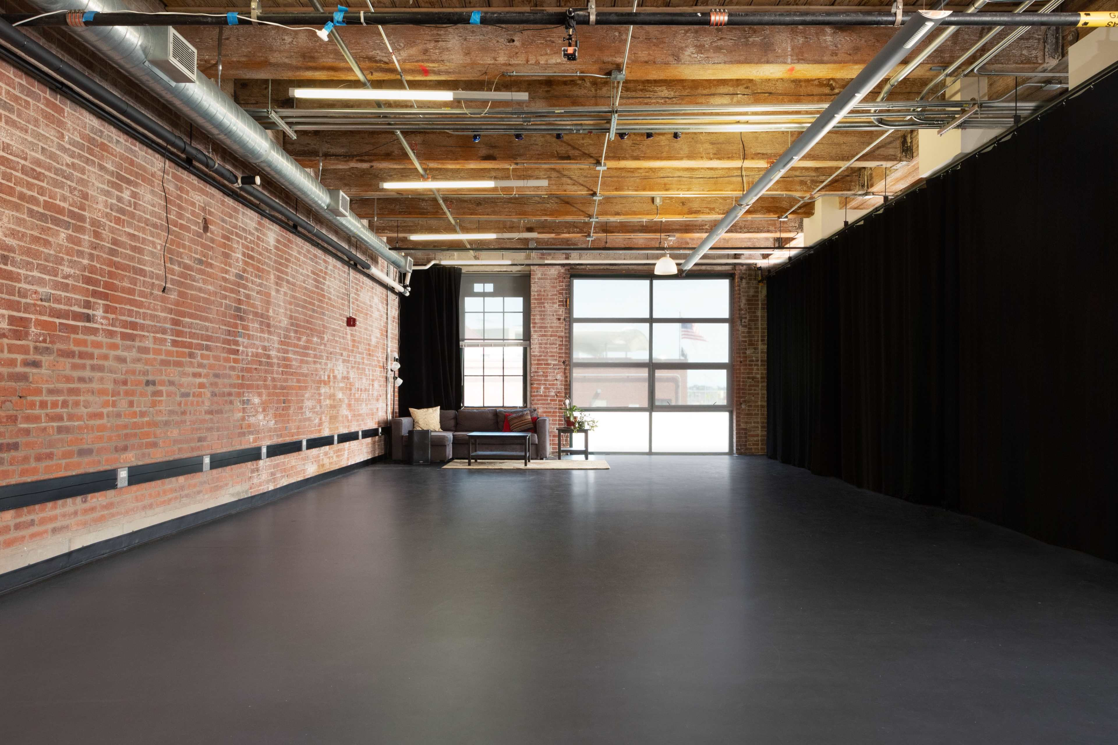 An industrial-style interior features exposed brick walls, wooden beams, and a dark floor, with a seating area in front of a large window.