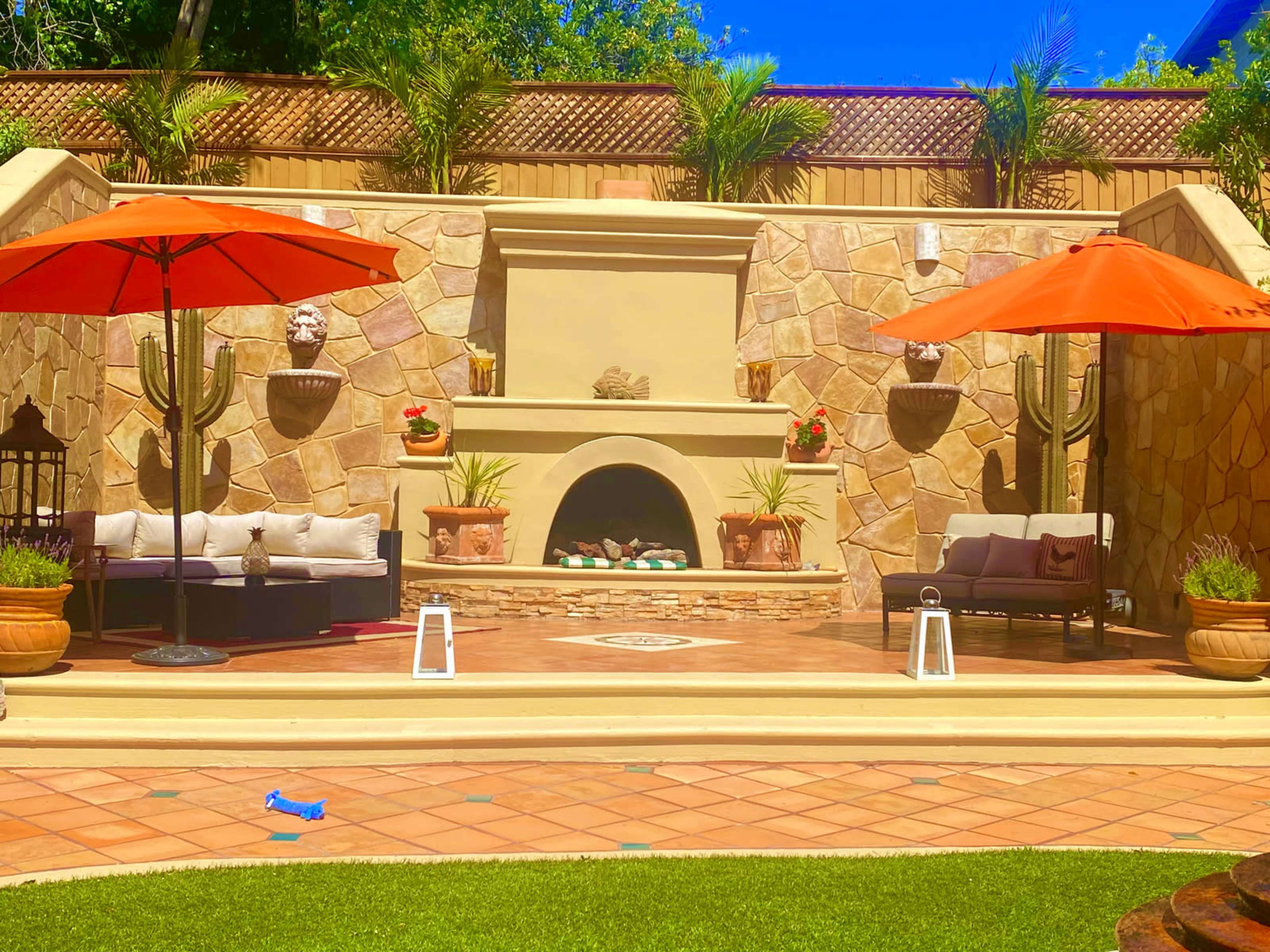 The outdoor patio features a stone wall with a fireplace and two red umbrellas shading seating areas, surrounded by potted plants and decorative accents.