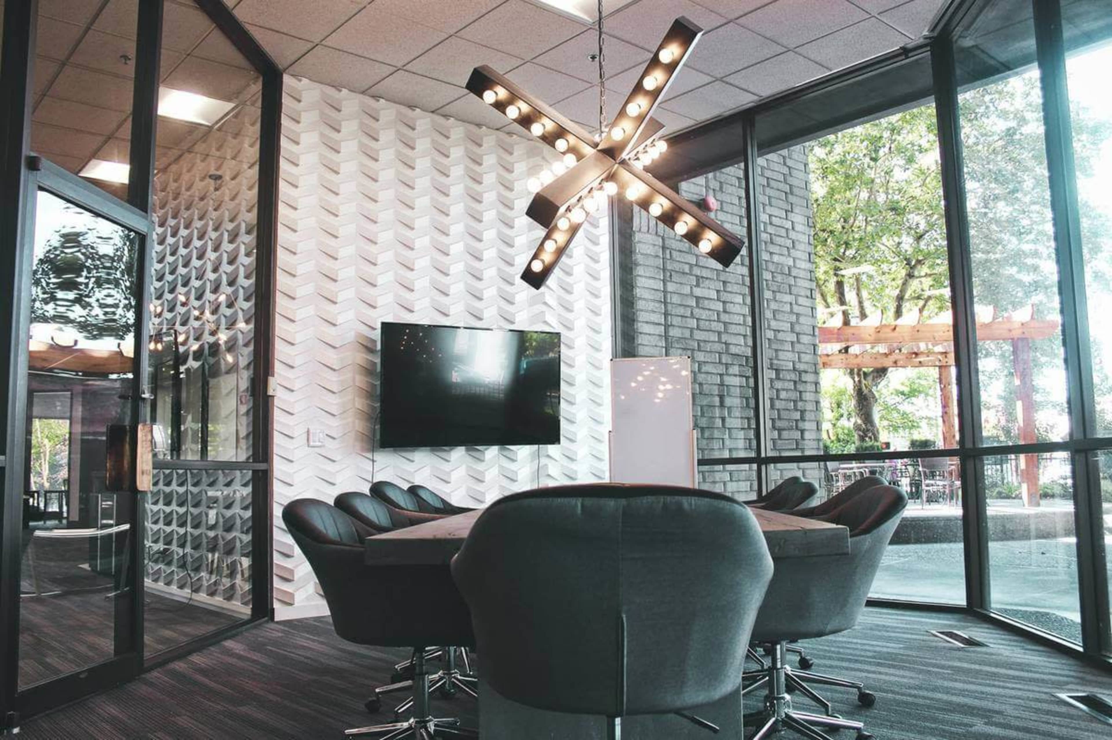 The image shows a modern conference room featuring a large table surrounded by chairs, a television on the wall, and a decorative light fixture above.