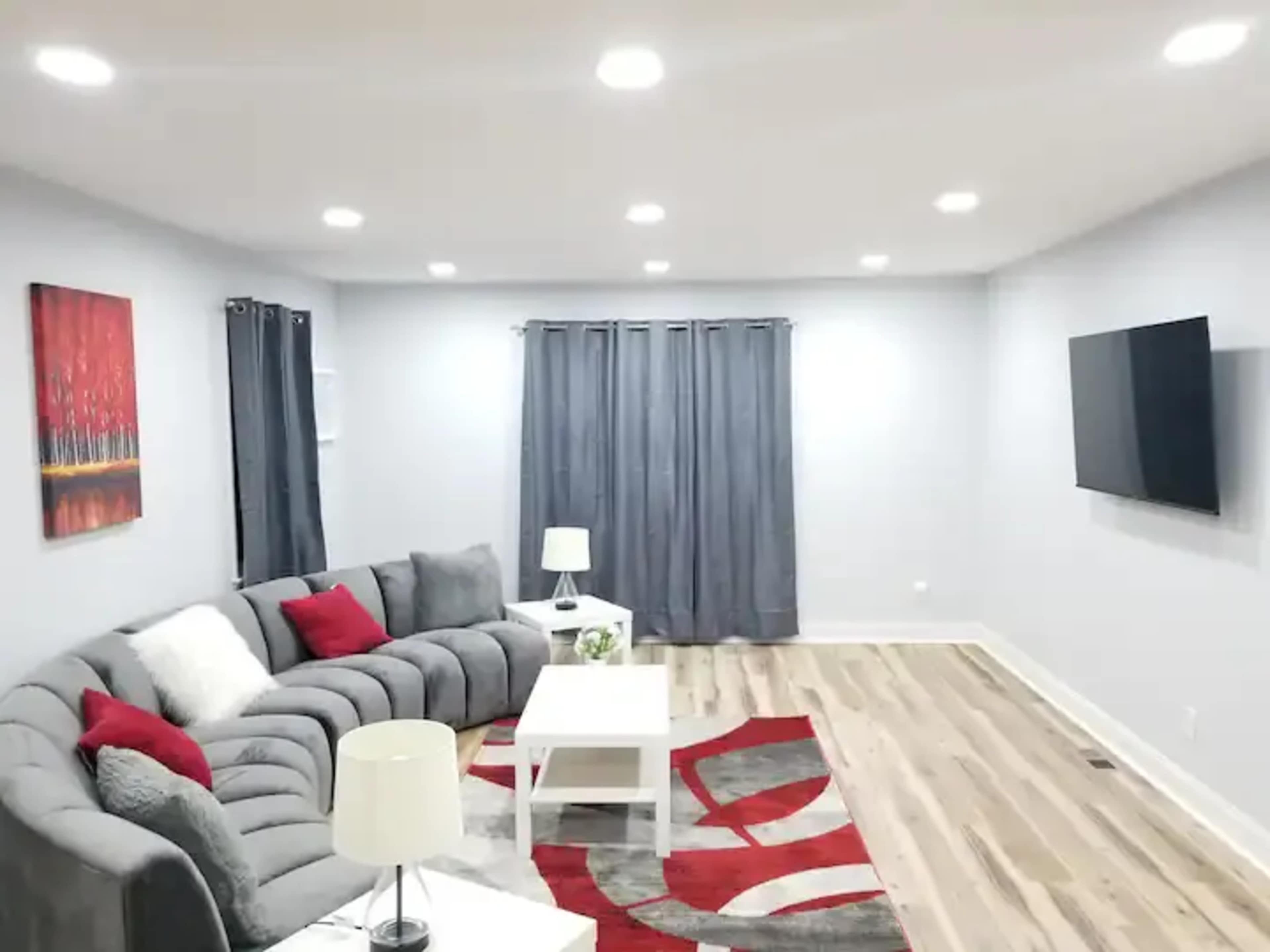 A modern living room features a gray sectional sofa, a white coffee table, a wall-mounted television, and decorative curtains, with a red and gray area rug on the wooden floor.