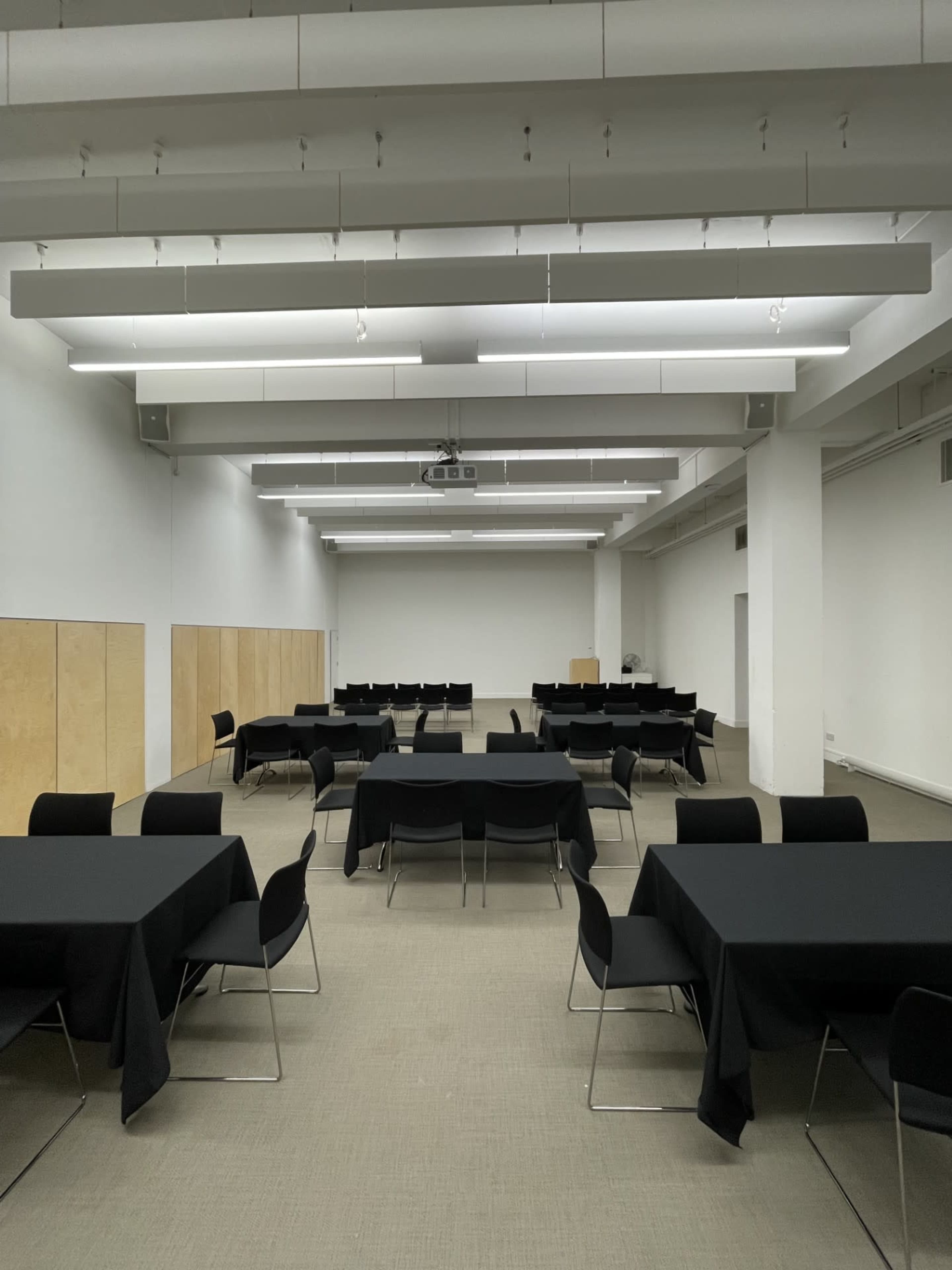 The image shows a spacious, empty conference room with multiple round tables covered in black tablecloths and chairs arranged around them, alongside wooden panels on the walls.
