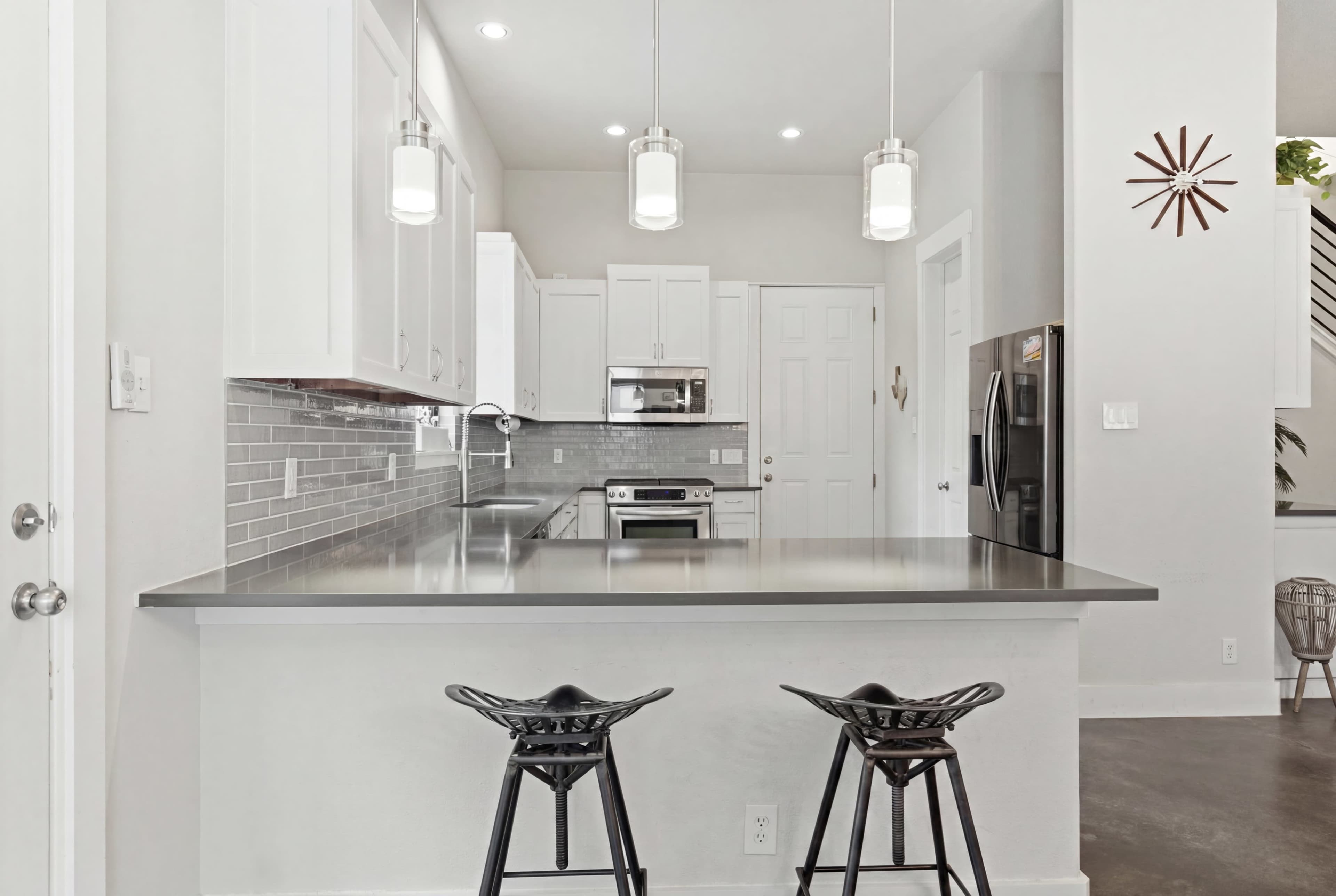 The image shows a modern kitchen with stainless steel appliances, white cabinetry, and two black bar stools positioned at a counter.