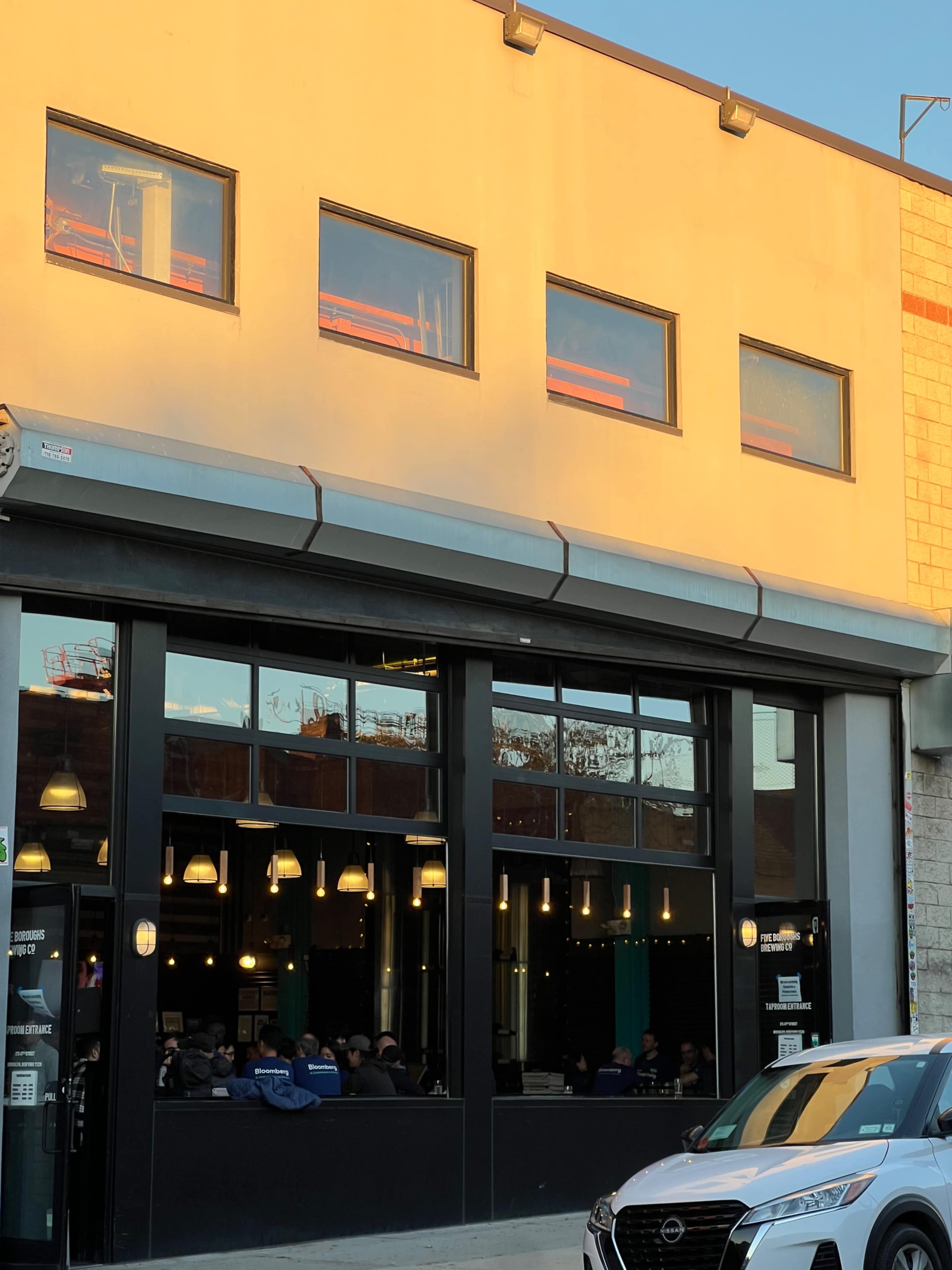 The image shows a modern building with large windows reflecting light, where a crowd is visible inside, and a parked car is in the foreground.