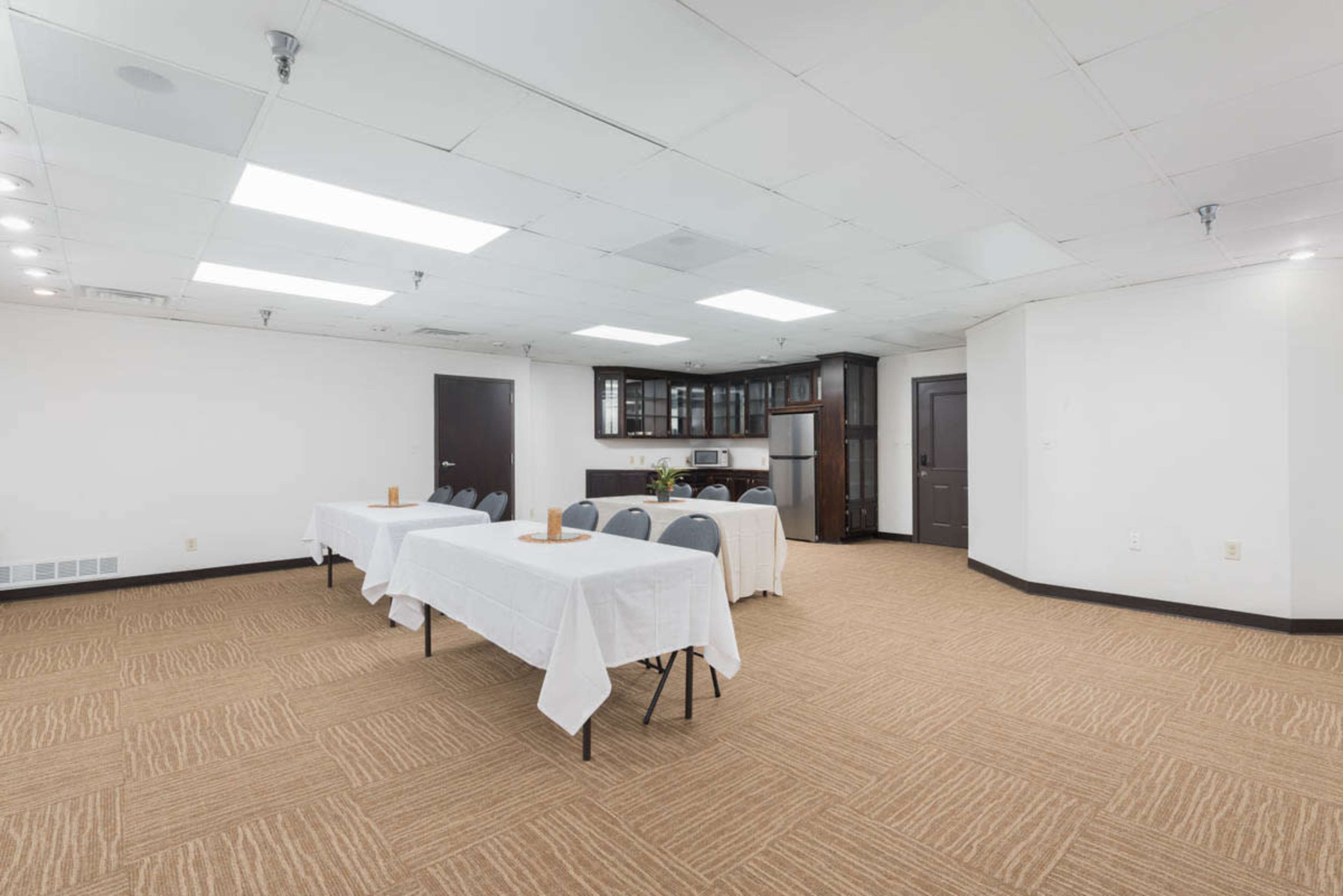 A brightly lit meeting room features multiple tables covered with white tablecloths arranged in a U-shape, with a kitchenette against the back wall.