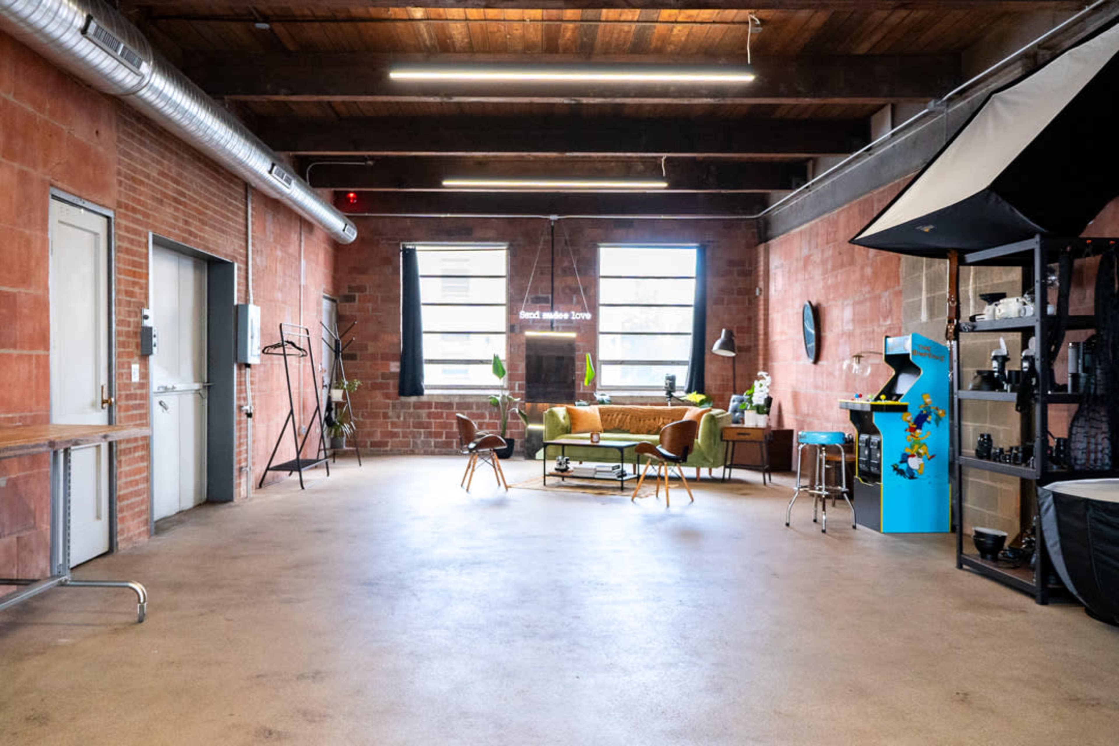 The image shows an interior space with exposed brick walls, wooden beams, a green sofa, and various furniture pieces arranged throughout.