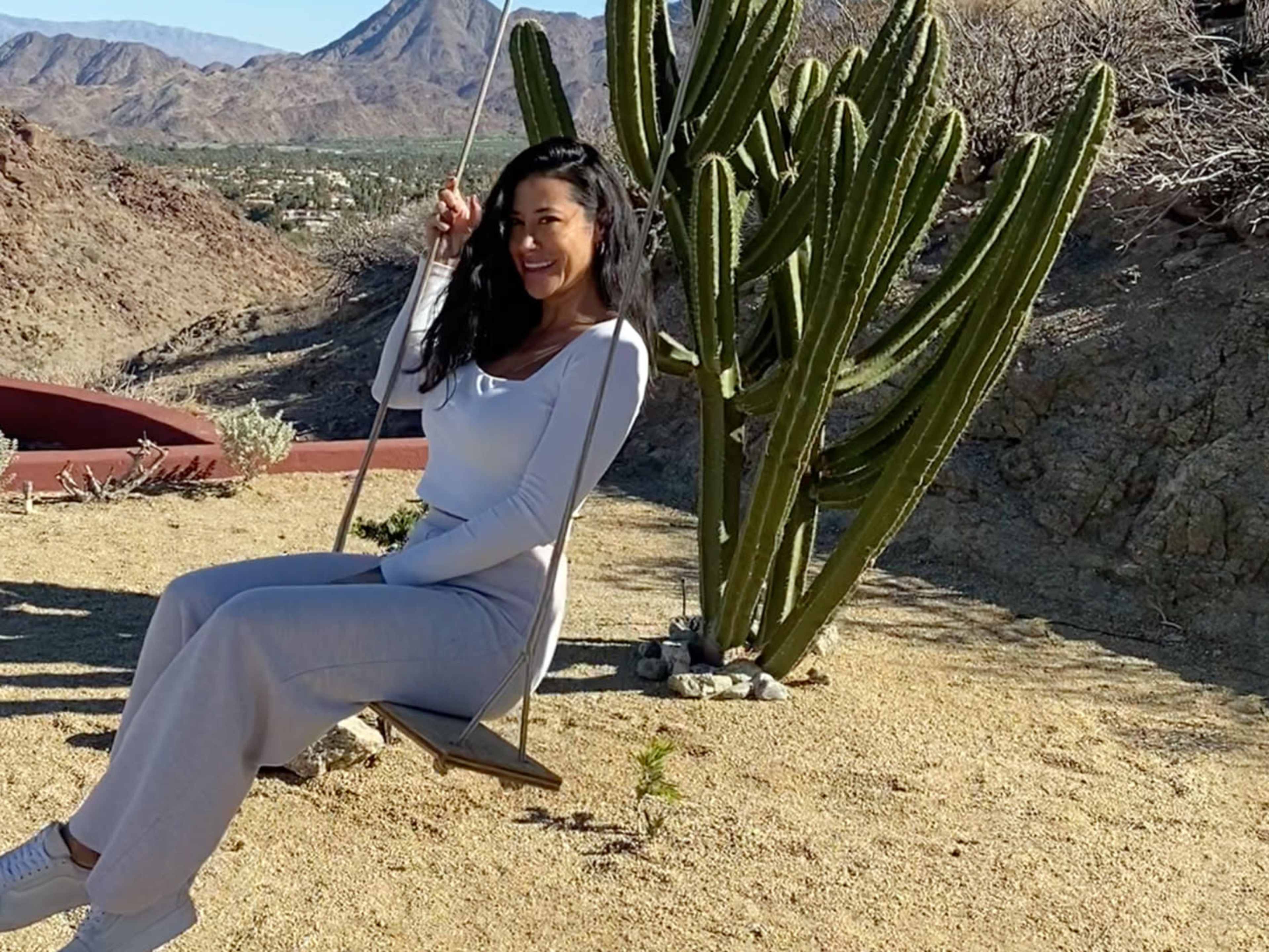 A woman in a white outfit sits on a swing near a tall cactus against a mountainous desert backdrop.
