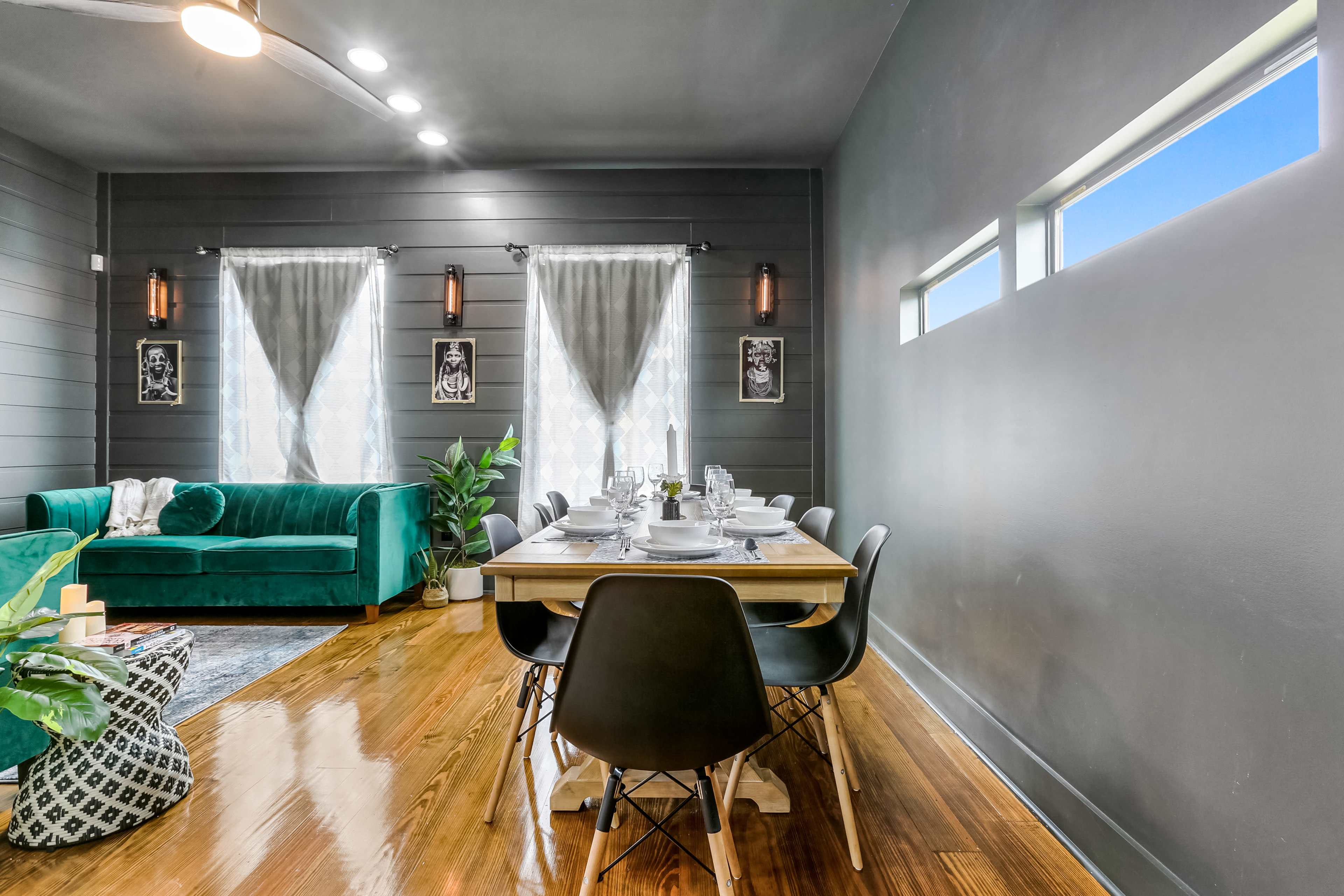 A dining area features a wooden table set for six, surrounded by black chairs, with a green velvet sofa and large windows in a softly lit gray room.