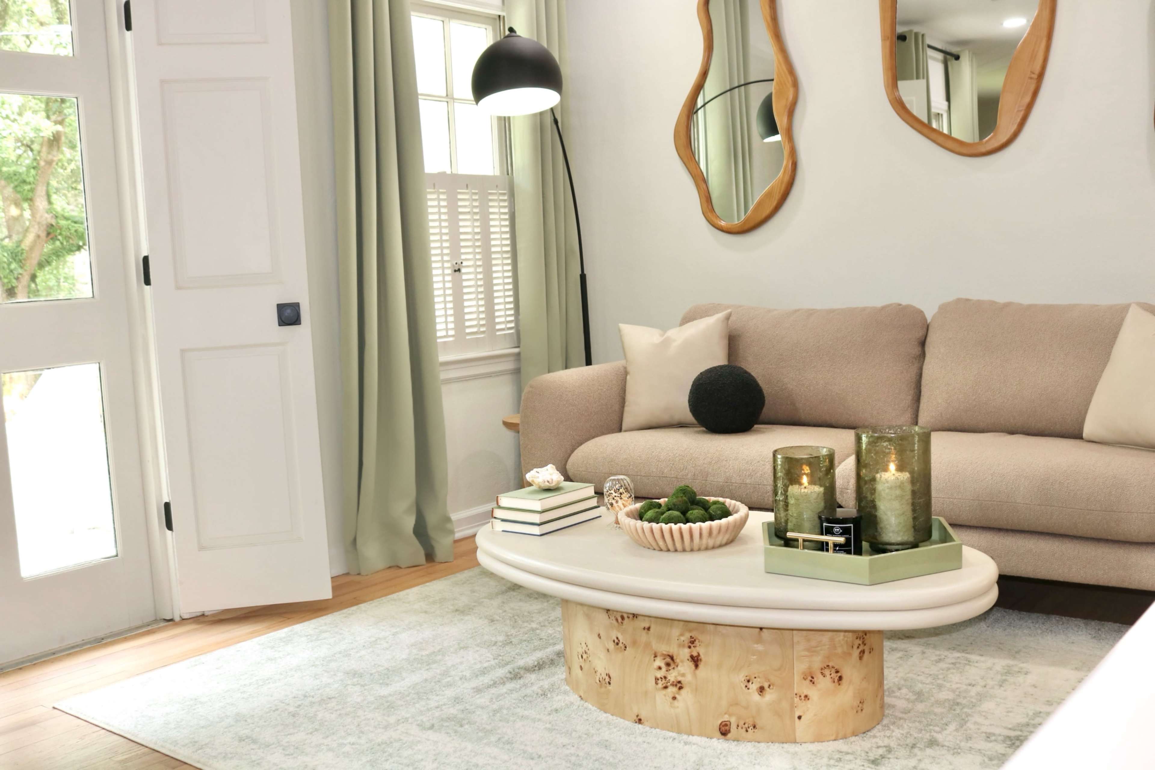 The image shows a cozy living room featuring a light-colored sofa, a circular coffee table with decorative items, and large windows with soft curtains.