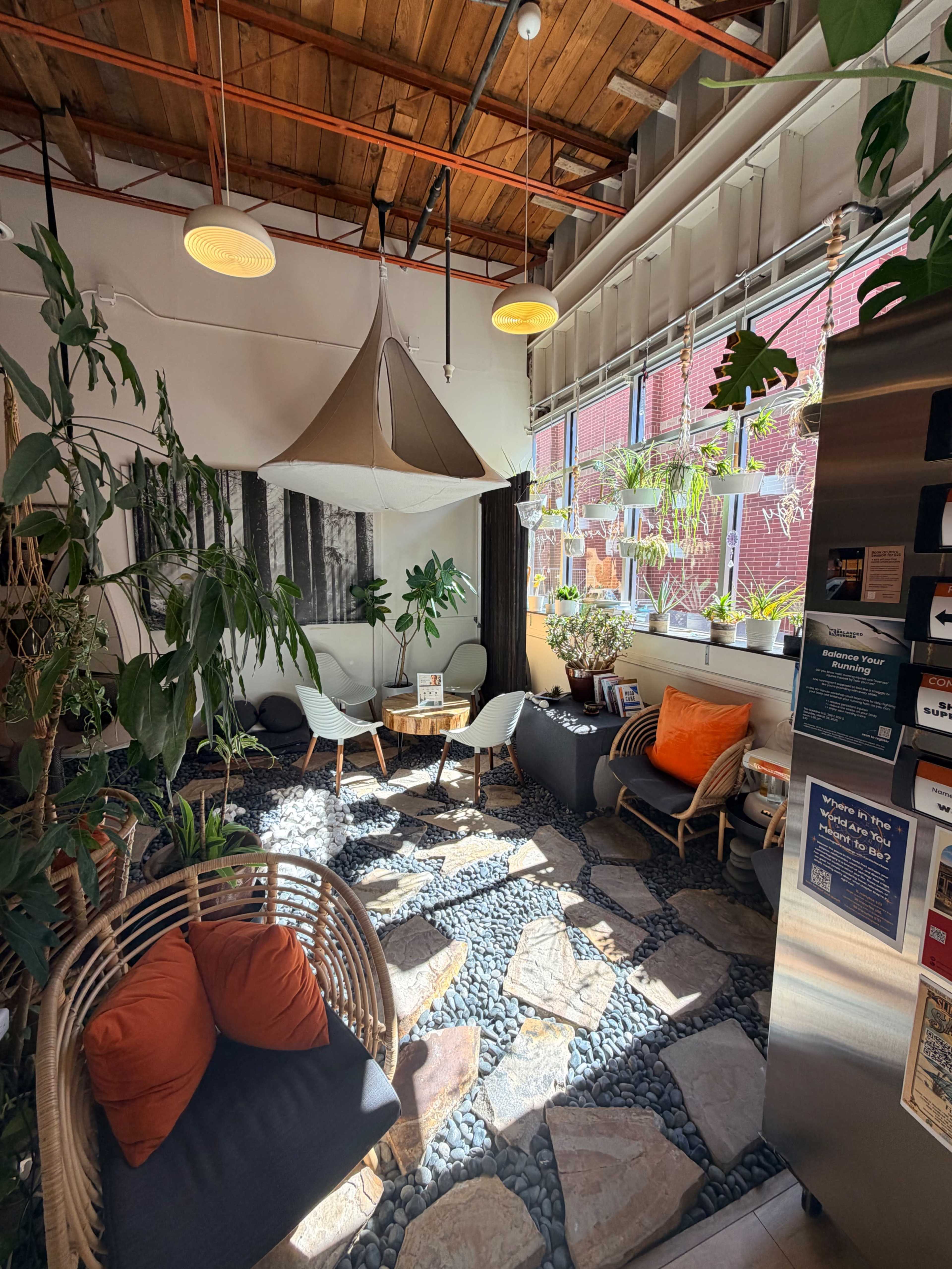 The image shows a cozy indoor seating area with a mix of plants, a stone and pebble floor, and modern furniture, illuminated by overhead lighting.