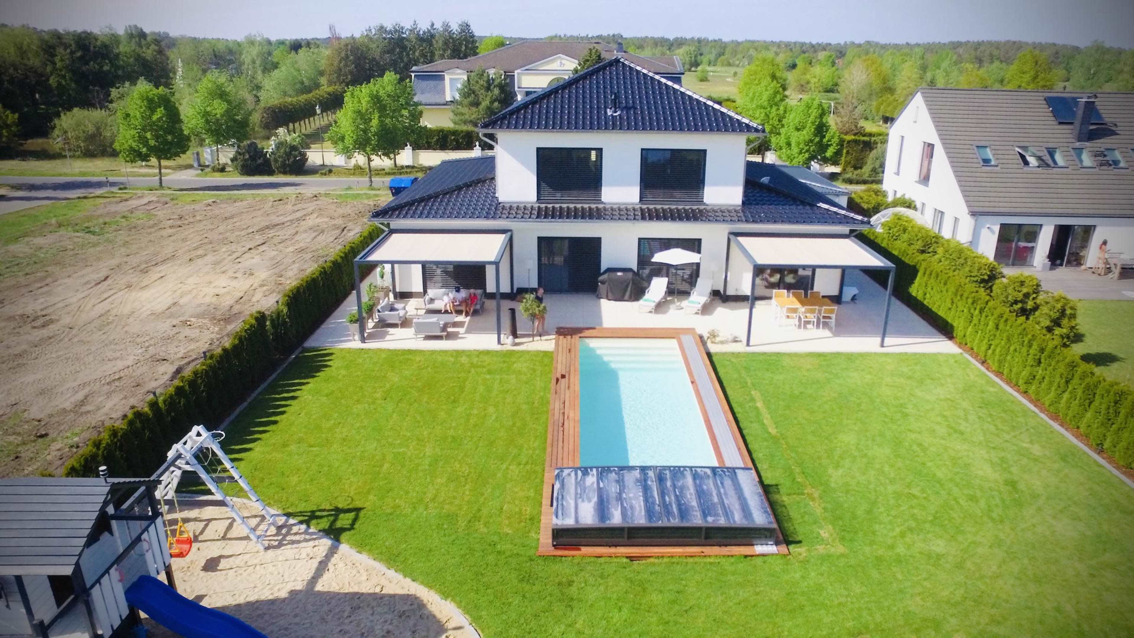 A modern two-story house with a pool and outdoor seating area is surrounded by a grassy lawn and trees.