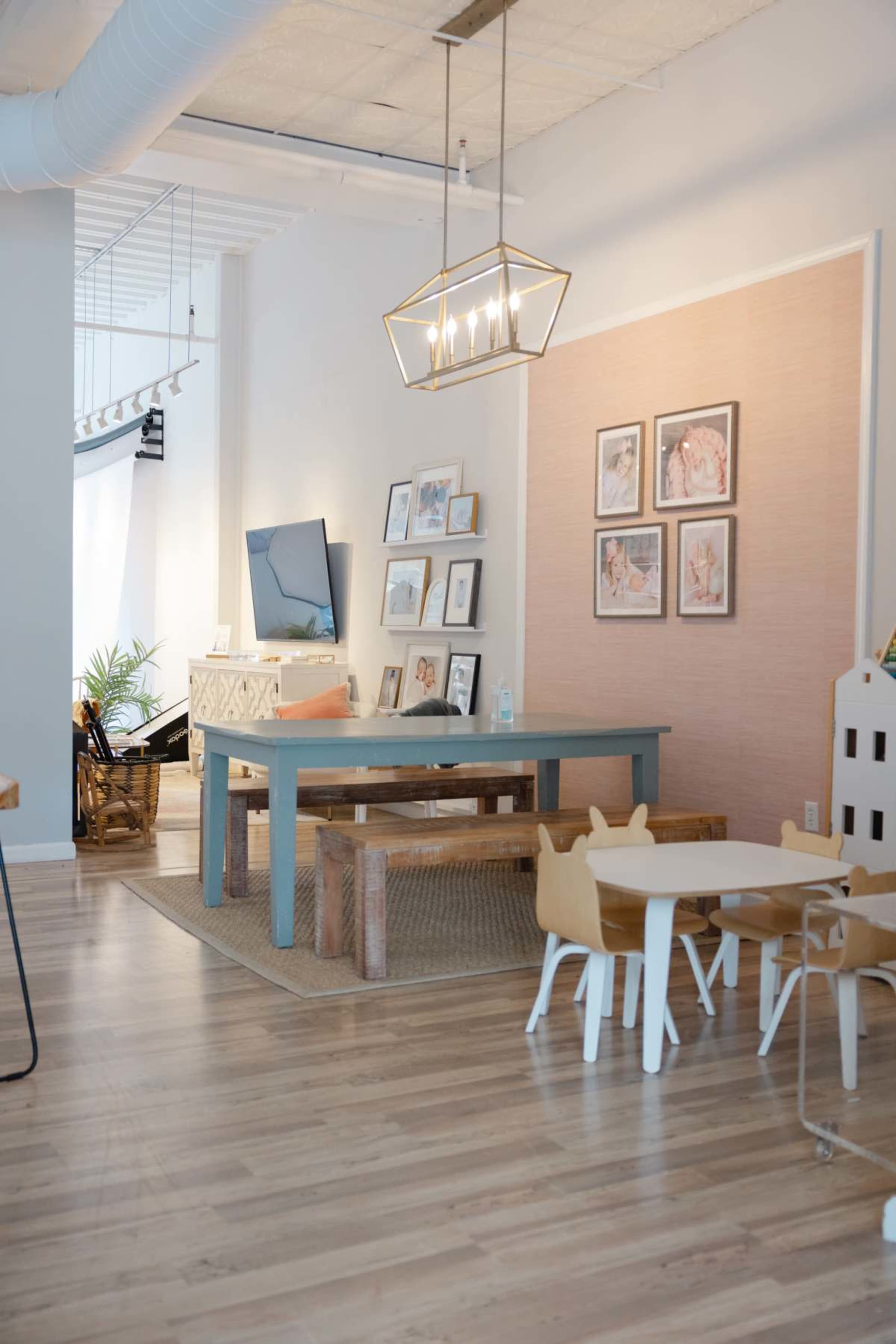 The image shows a modern, spacious interior featuring a dining table with benches, a television mounted on the wall, and framed artwork arranged nearby.