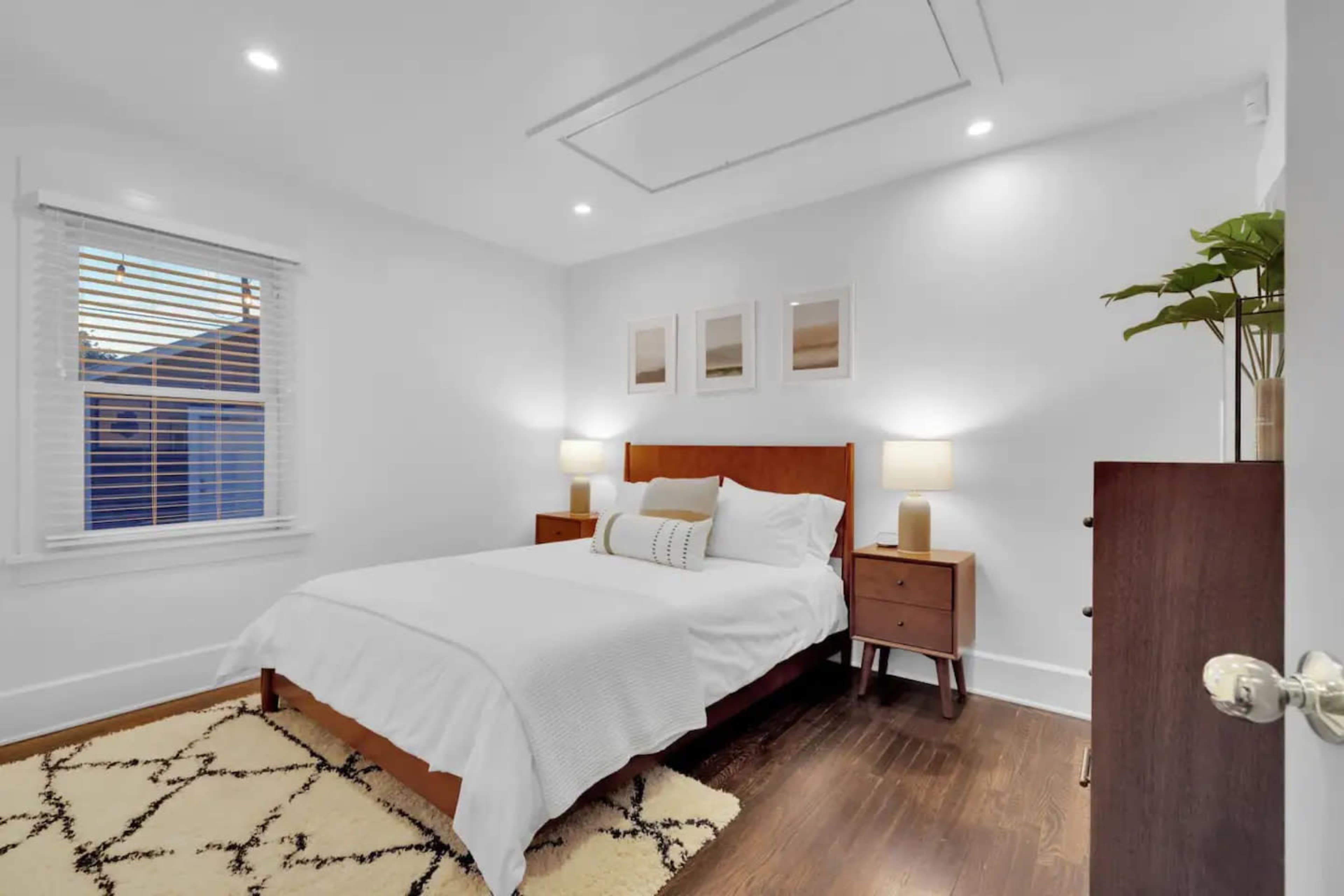 The image shows a neatly arranged bedroom featuring a bed with white bedding, two nightstands with lamps, and a dresser, all set against a white wall with framed pictures and a plant.