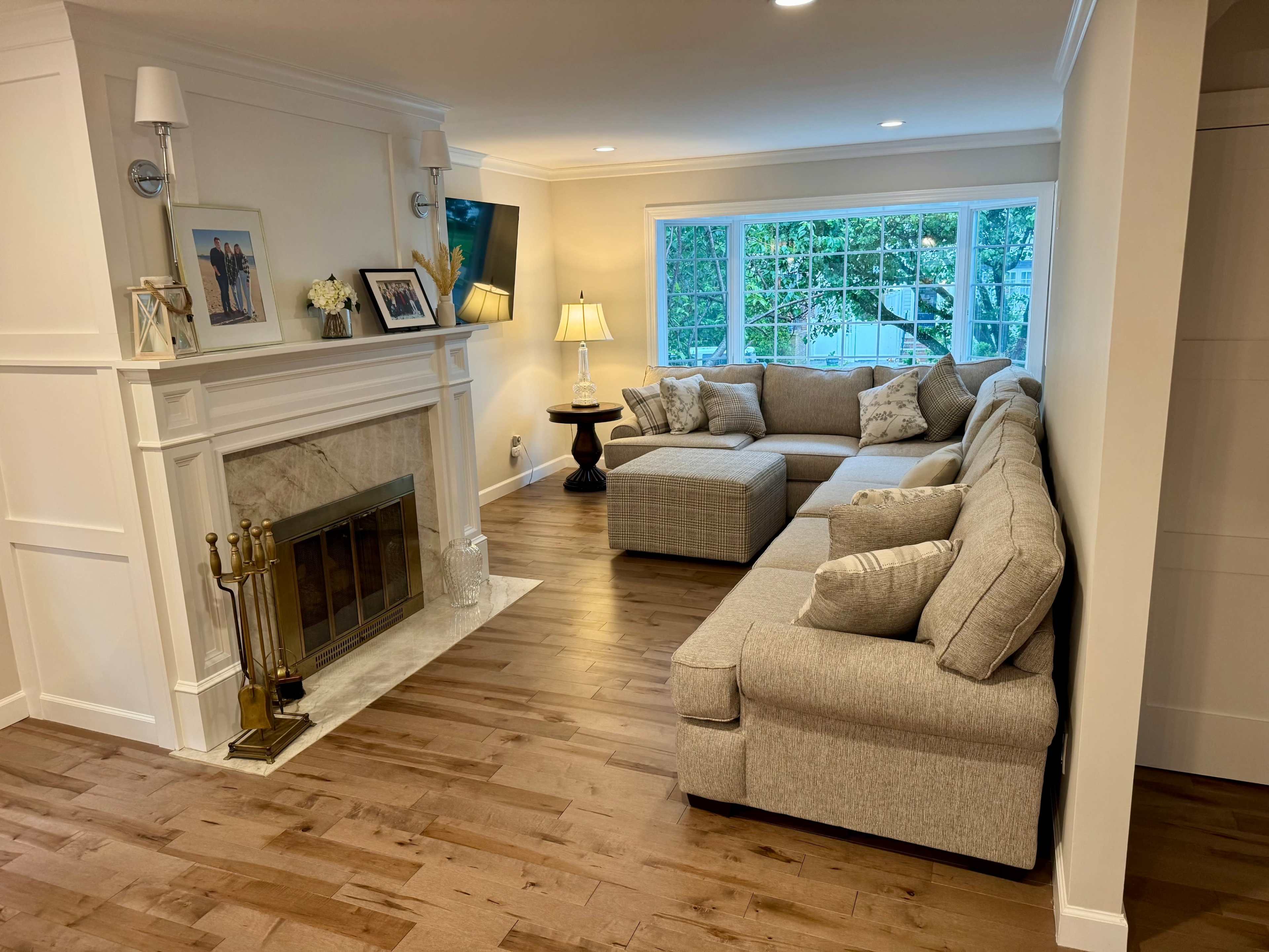 The image shows a living room with a sectional sofa, a coffee table, a fireplace, and a large window allowing natural light to enter.