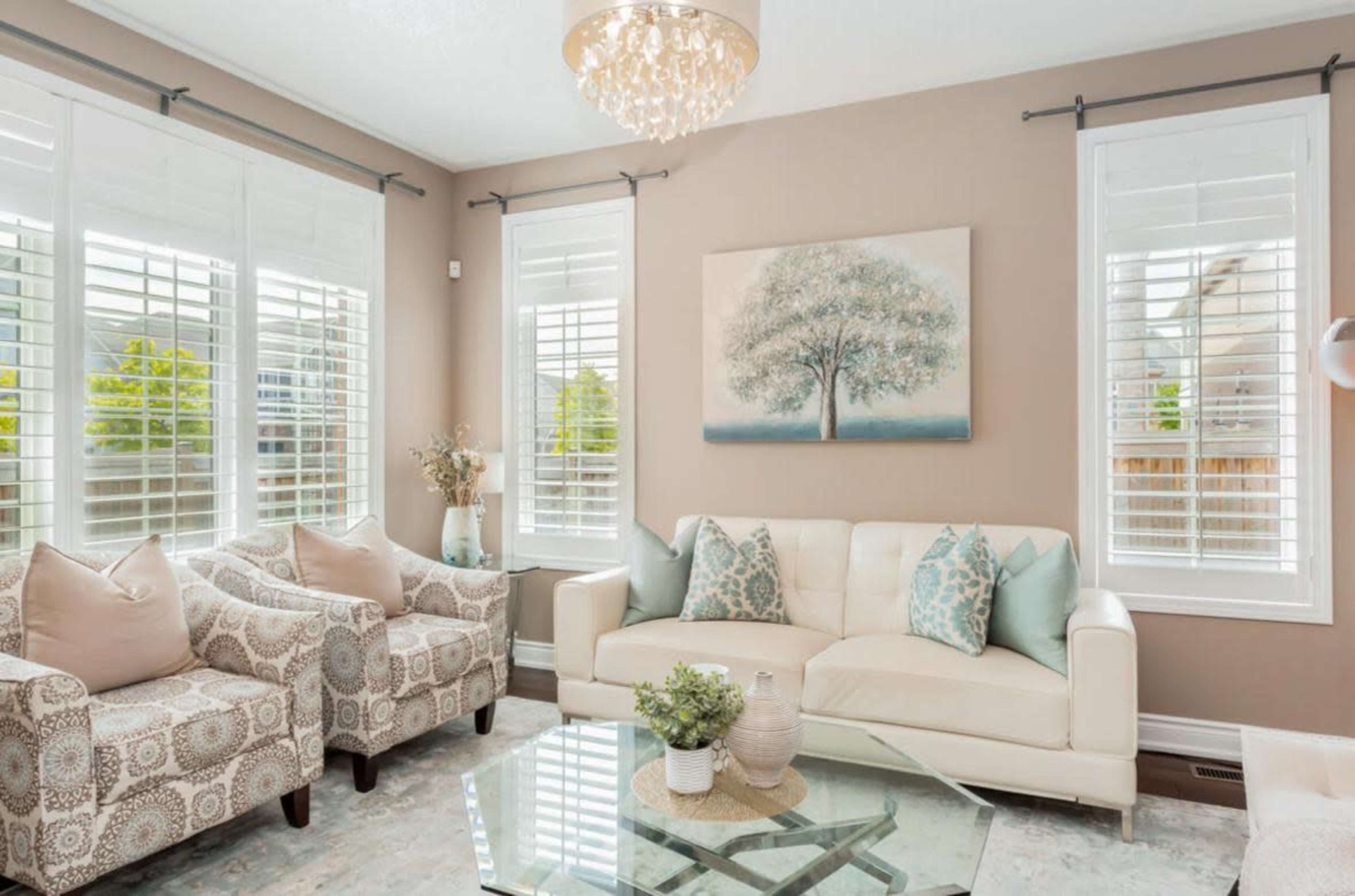 The image features a well-lit living room with a light-colored sofa, two patterned armchairs, and a glass coffee table, surrounded by large windows with white shutters.