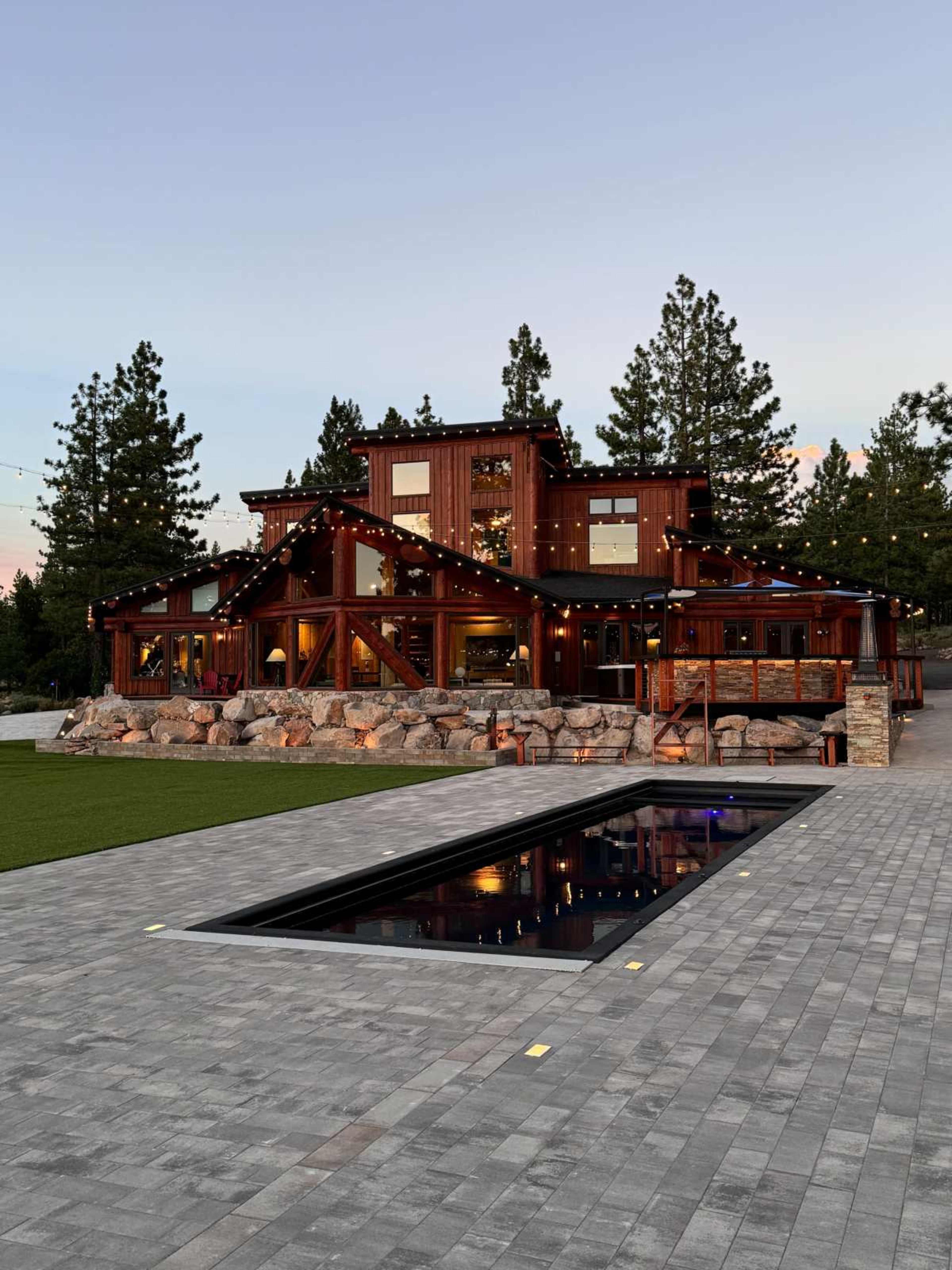A large wooden house with a modern design stands next to a rectangular swimming pool surrounded by stone and grass, framed by tall trees in the background.