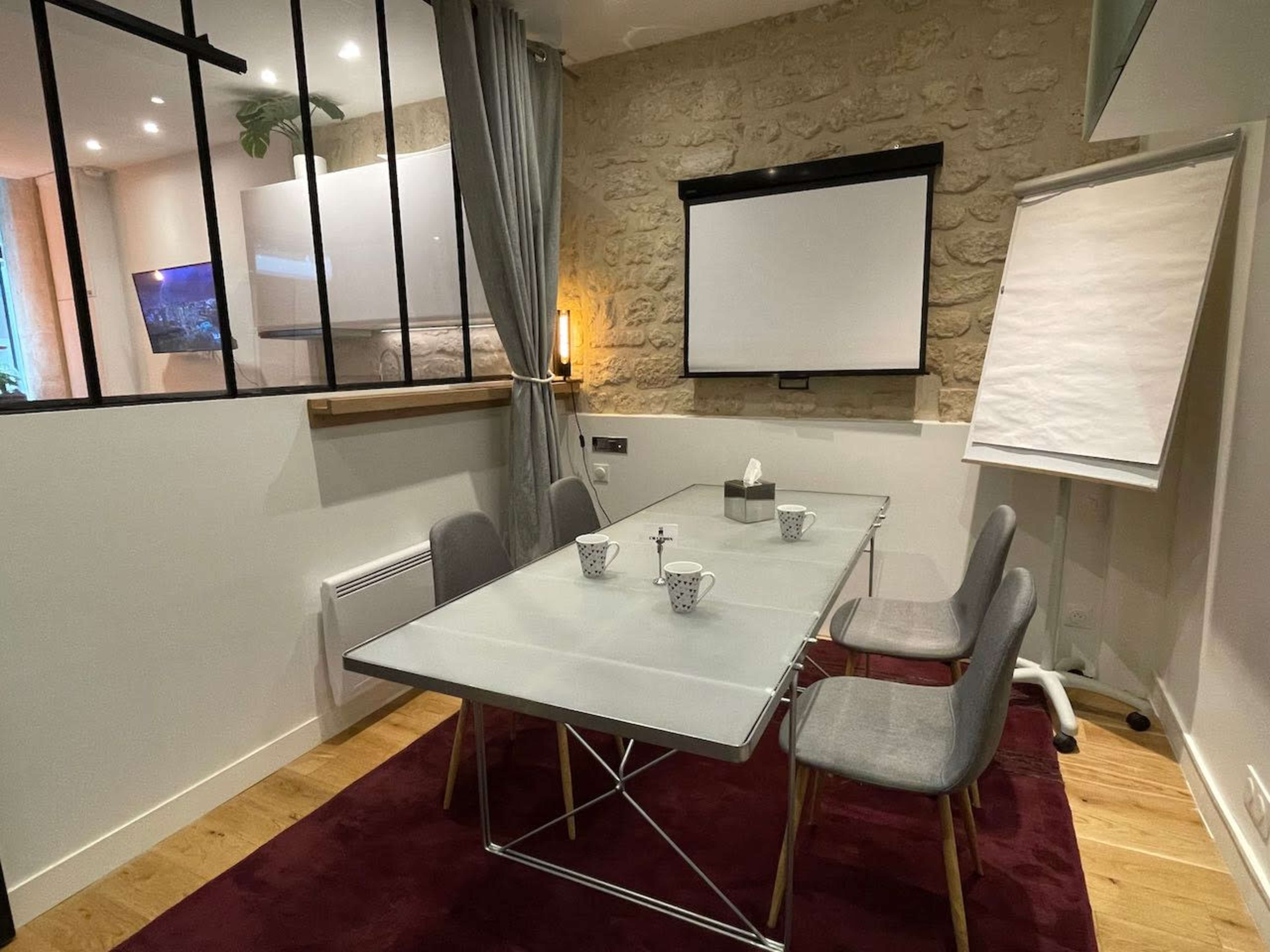 A glass-top conference table with four gray chairs is set up in a room with stone walls and a projector screen at one end.
