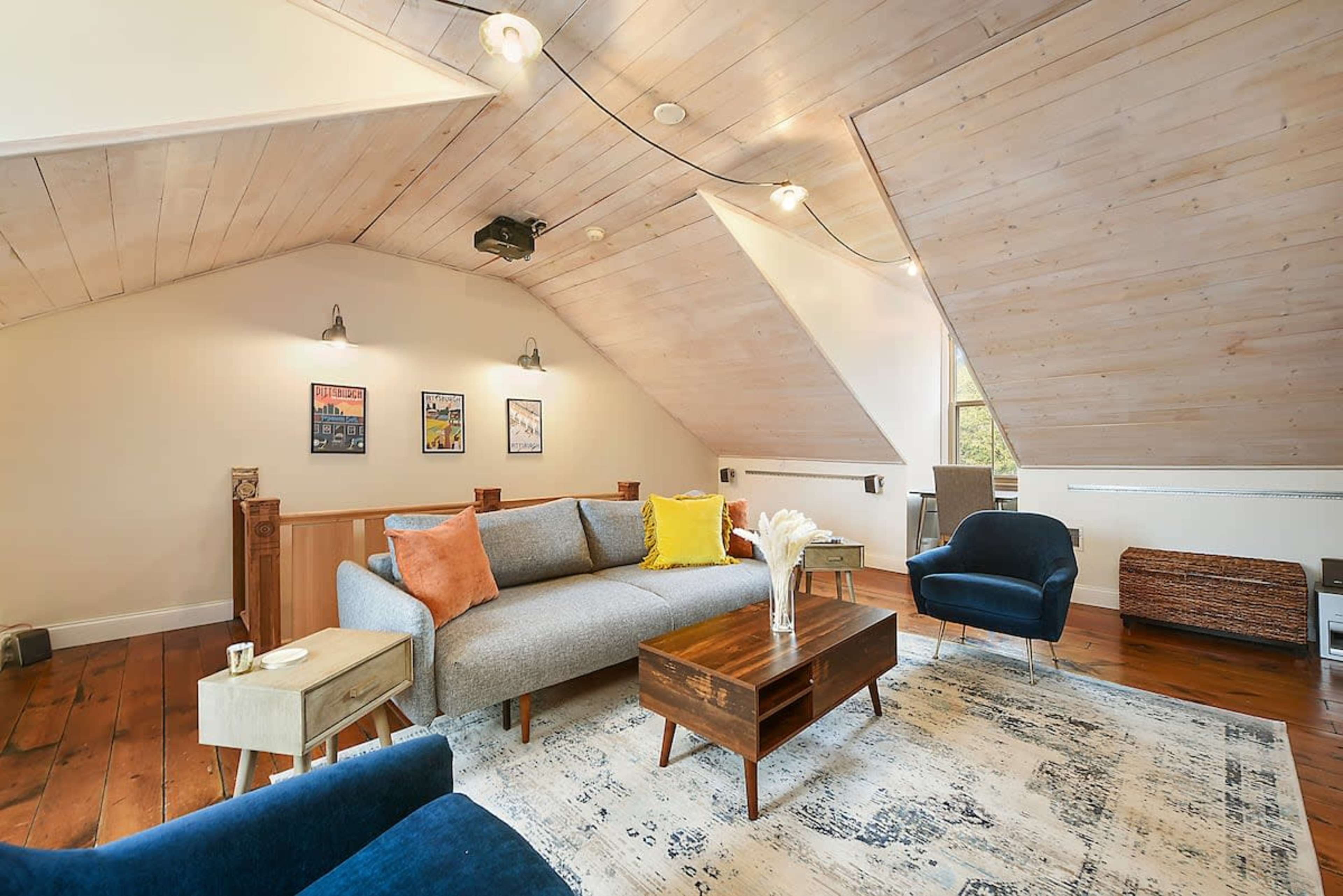 The image shows a cozy attic living room featuring a gray sofa, two accent chairs, a wooden coffee table, and framed artwork on the walls.