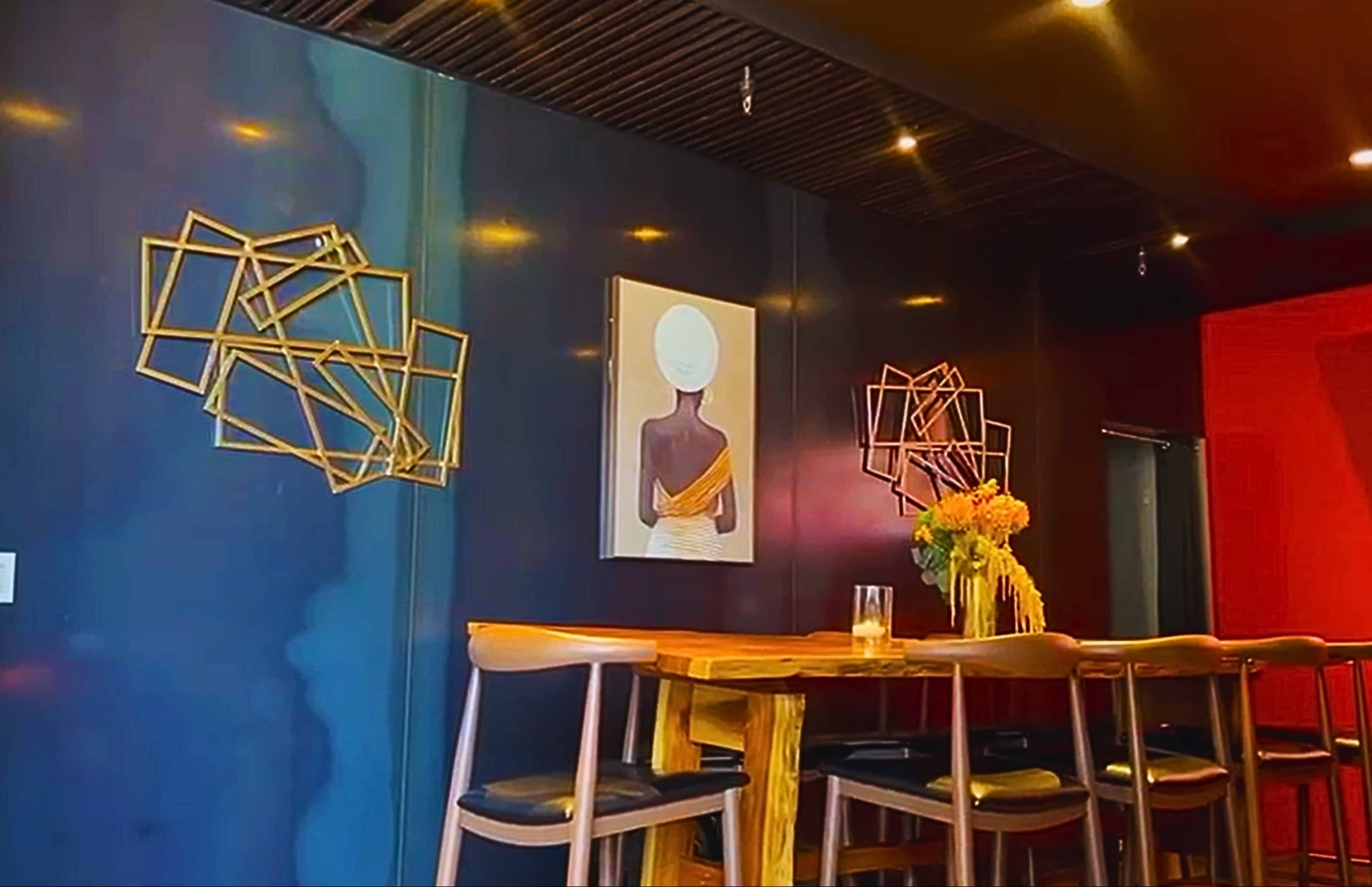 The image shows a modern interior with a dark wall featuring abstract gold decor, a framed painting of a woman, and a wooden bar with stools.