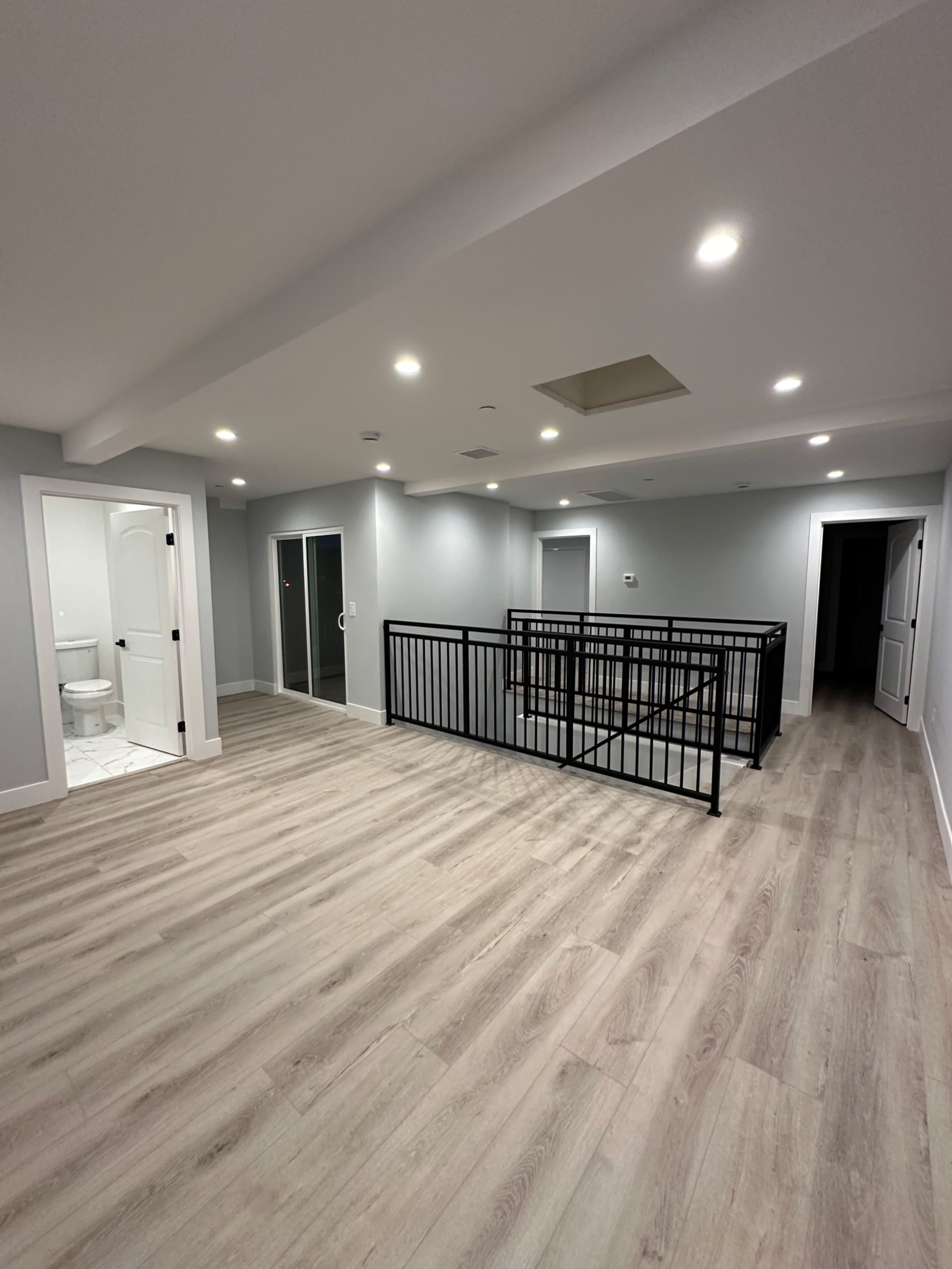 The image shows a spacious, modern interior room with light wood flooring, gray walls, and a black railing around a central area, with a bathroom visible in the background.