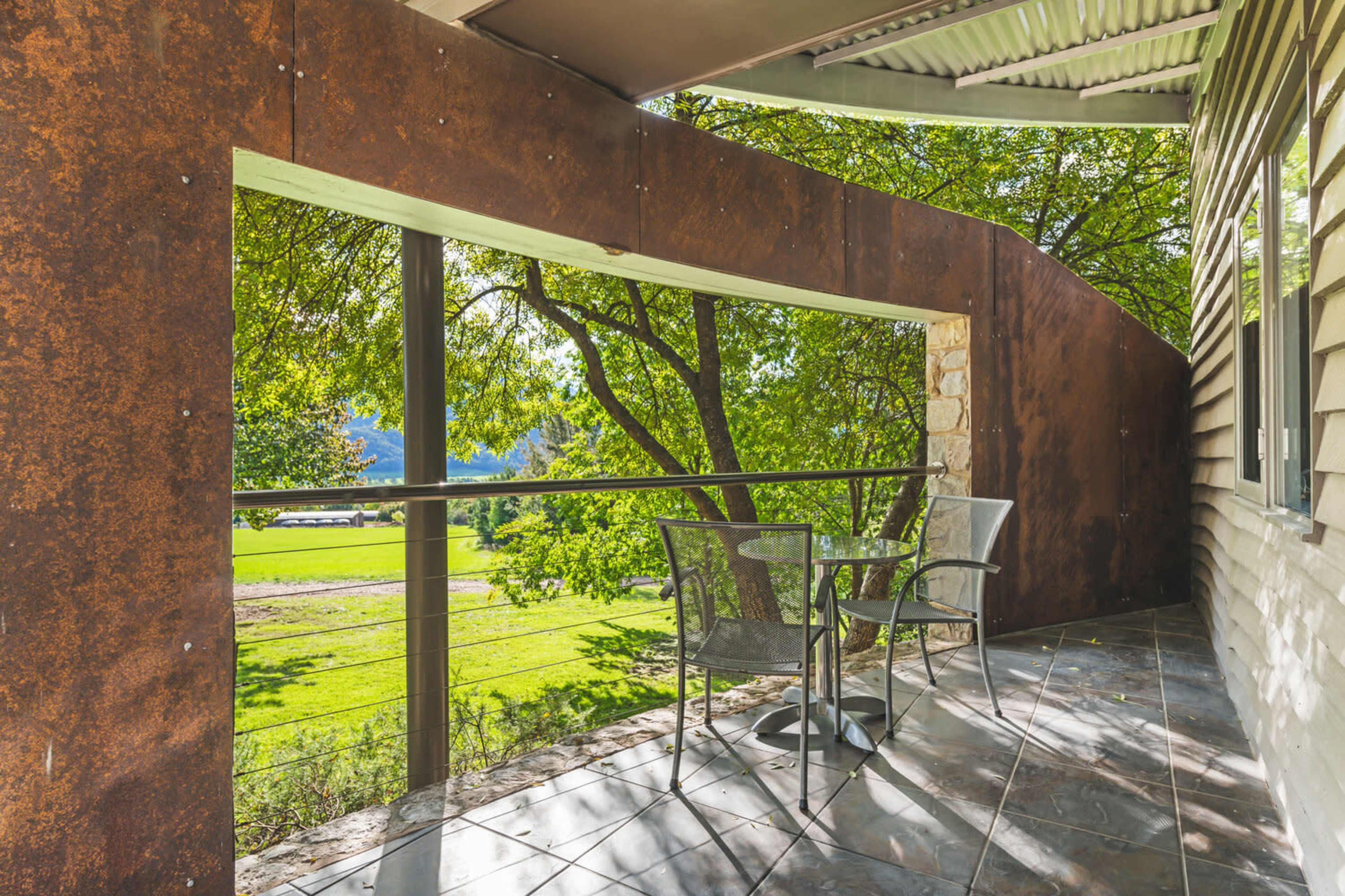 A balcony with metal railings and two chairs overlooking a green landscape through an open window.
