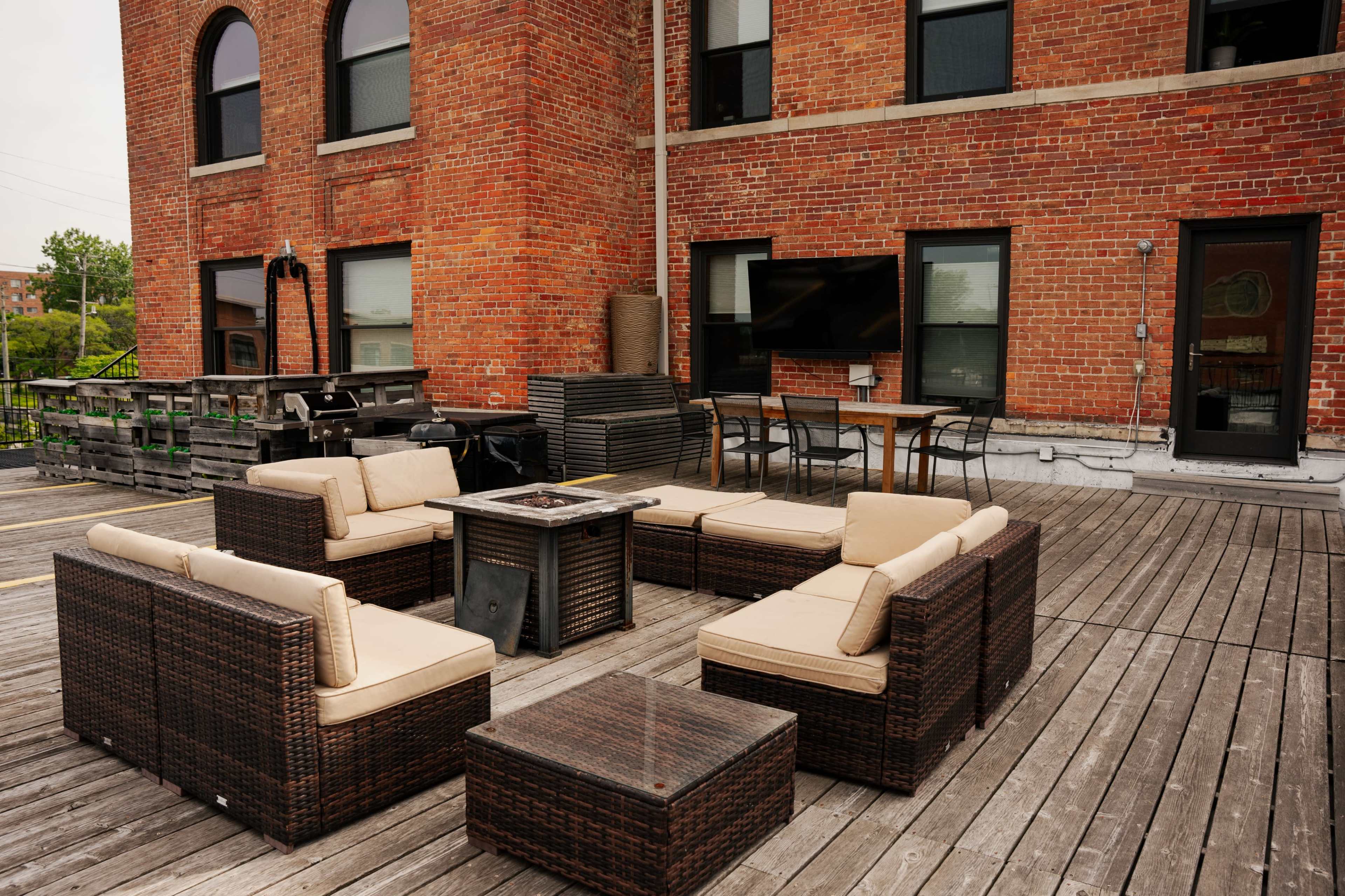 The image shows a patio area with a circular arrangement of wicker seating and a fire pit, complemented by a dining table and chairs against a brick building.