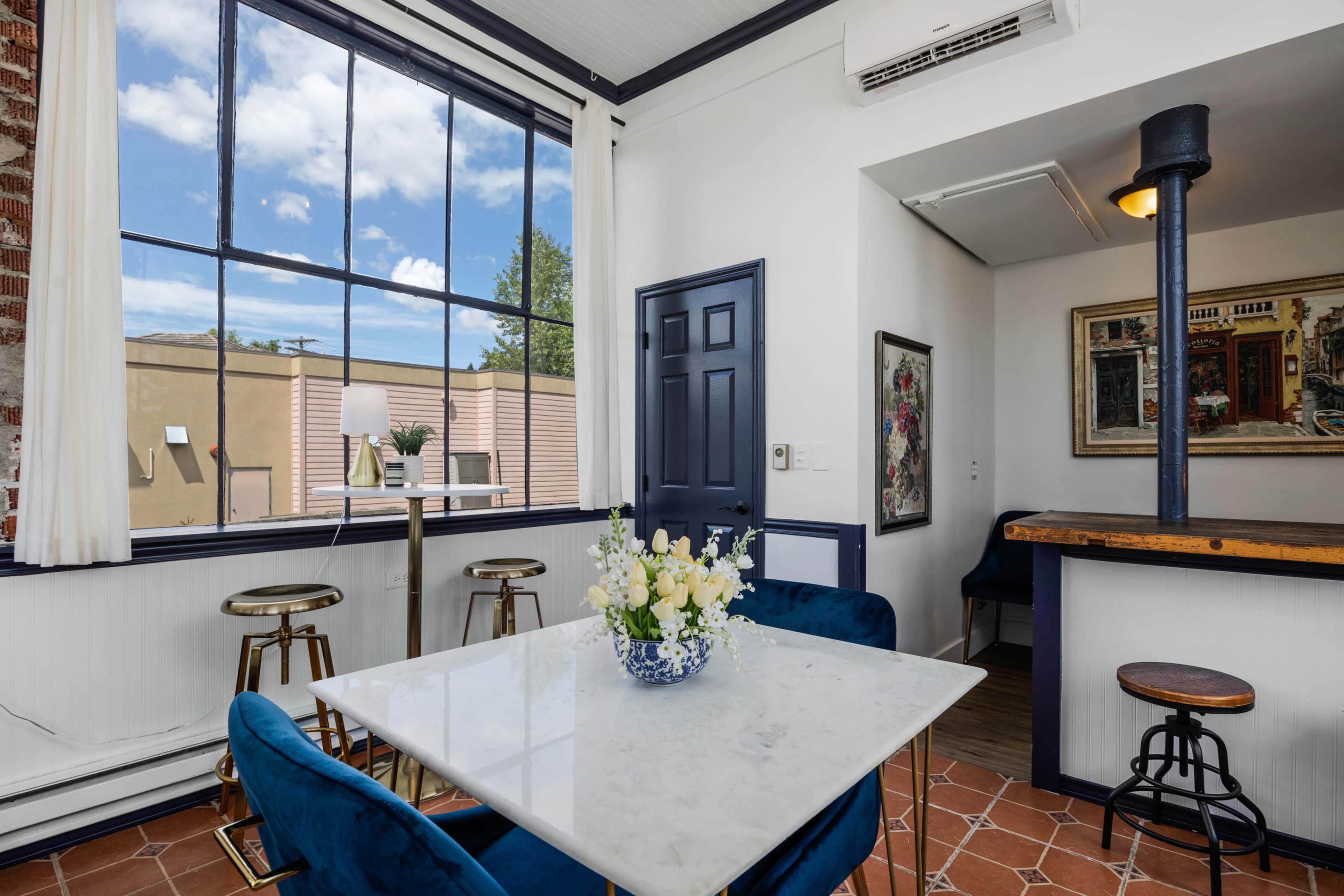 A dining area features a marble table surrounded by blue chairs, with large windows allowing natural light and showcasing the outdoor view.