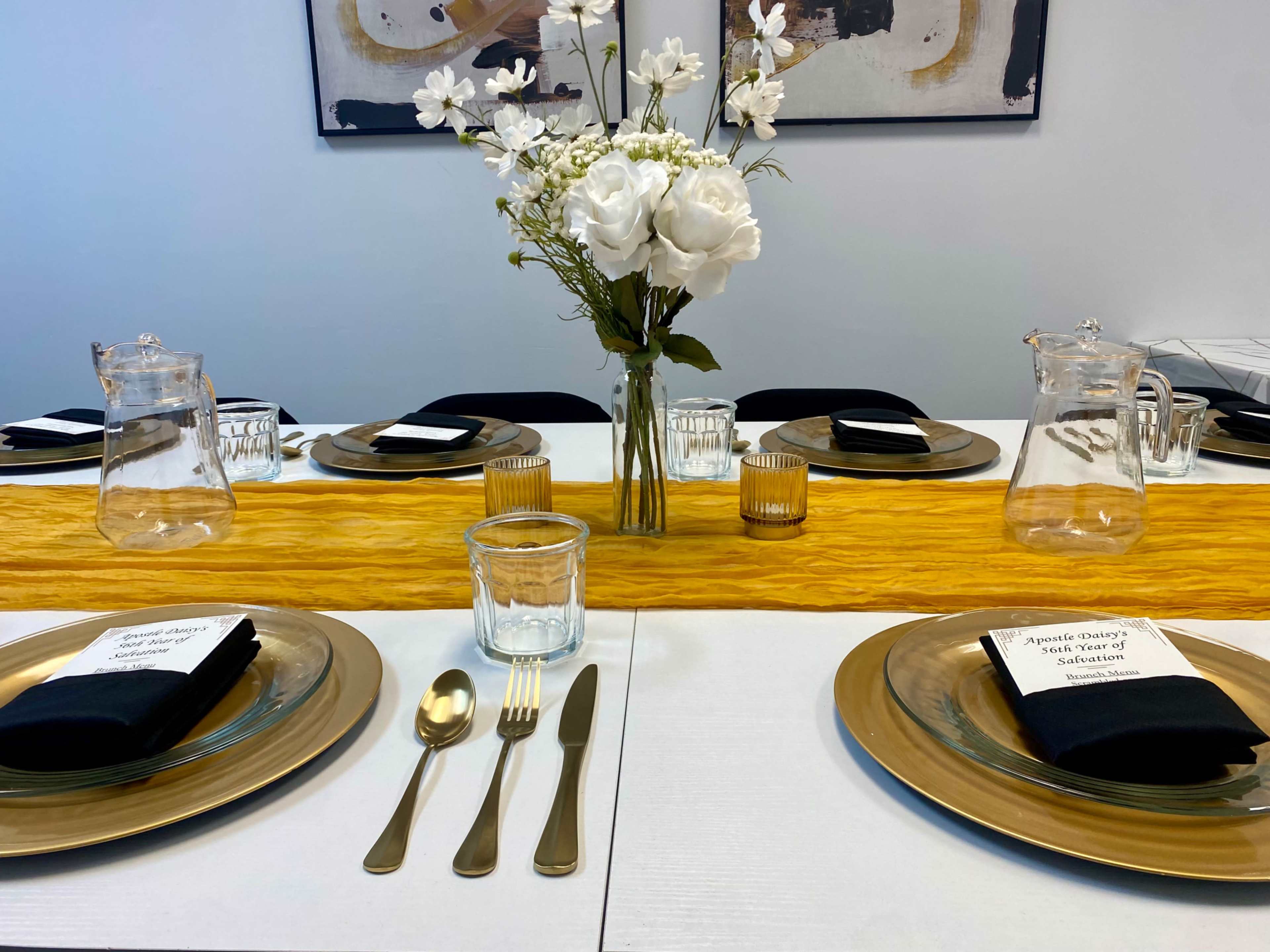 A dining table is set with gold plates, black napkins, glassware, and a yellow table runner, topped with a vase of white flowers.
