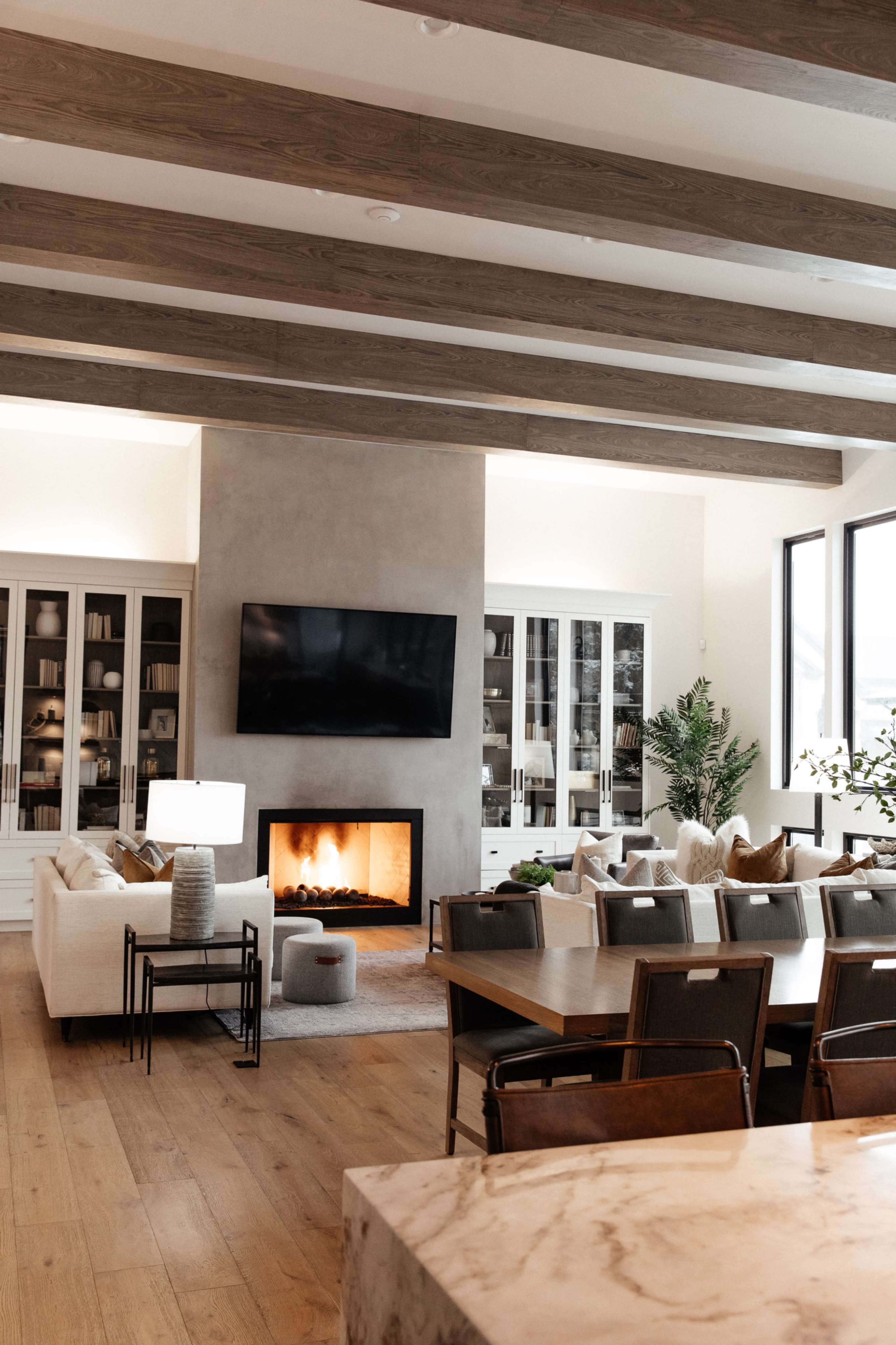 A modern living room featuring a fireplace, large windows, wooden beams on the ceiling, and a mix of comfortable seating arrangements.